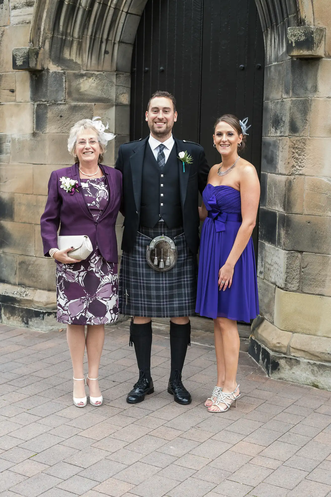 Three people posing for a photo at a wedding, featuring a man in a kilt flanked by two women in elegant dresses, standing in front of an arched stone doorway.