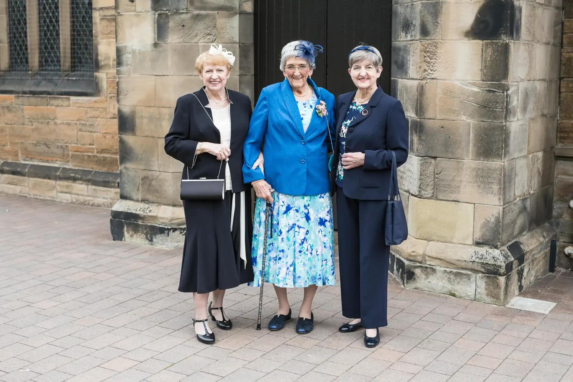 Three elderly women smiling and standing together outside a stone building, dressed in formal attire with floral accents.