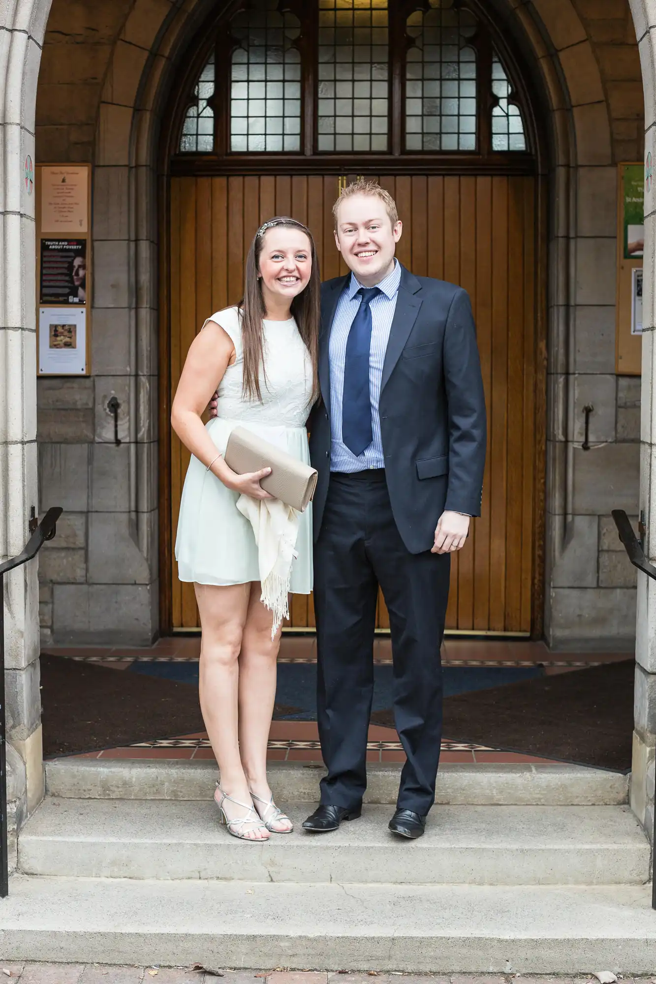 A smiling couple standing in front of a church entrance, the woman holding a book and the man in a suit.