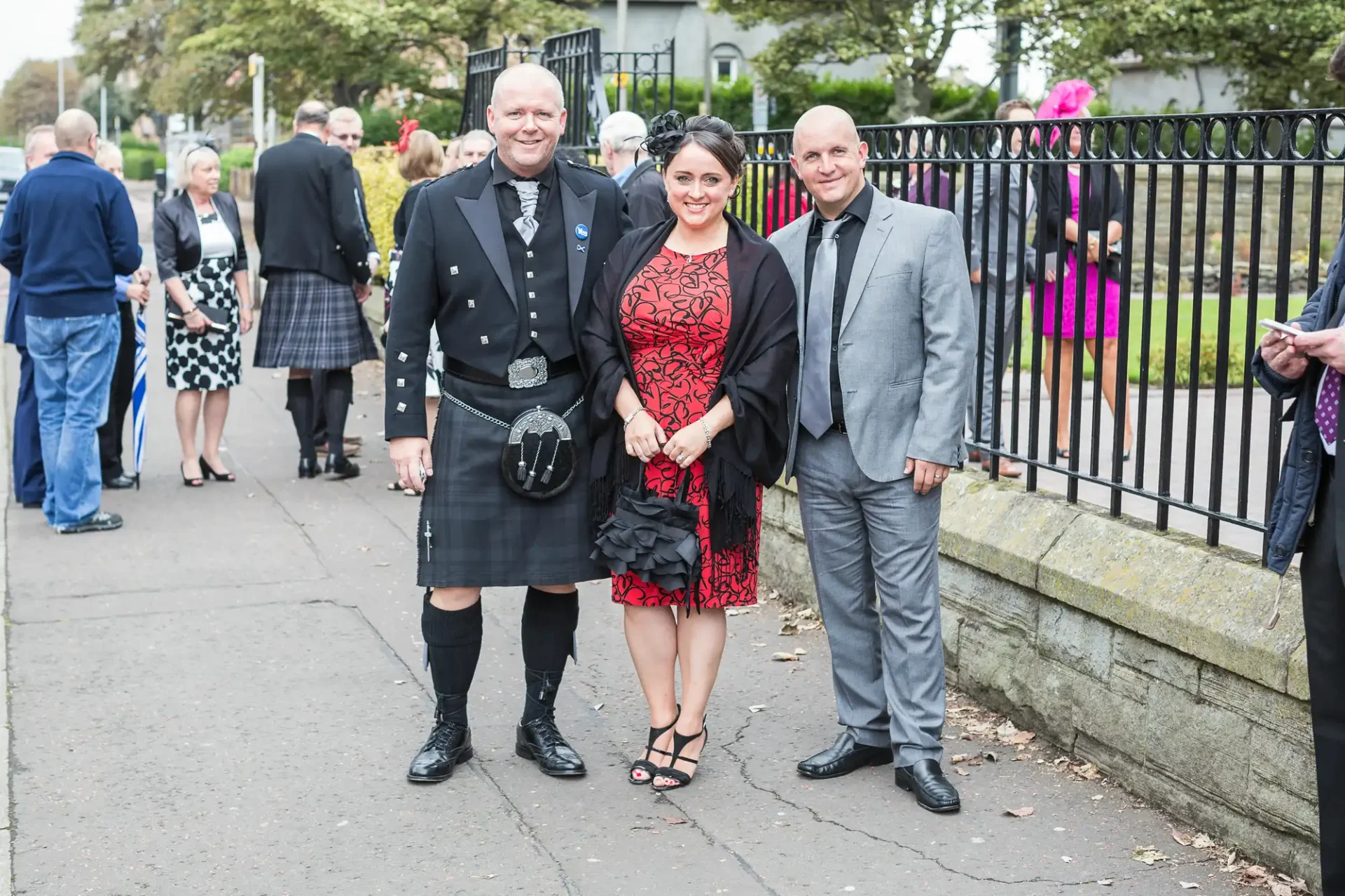 Two men and a woman posing for a photo at a social gathering, one man in a kilt and the other two in formal wear, with other guests in the background.