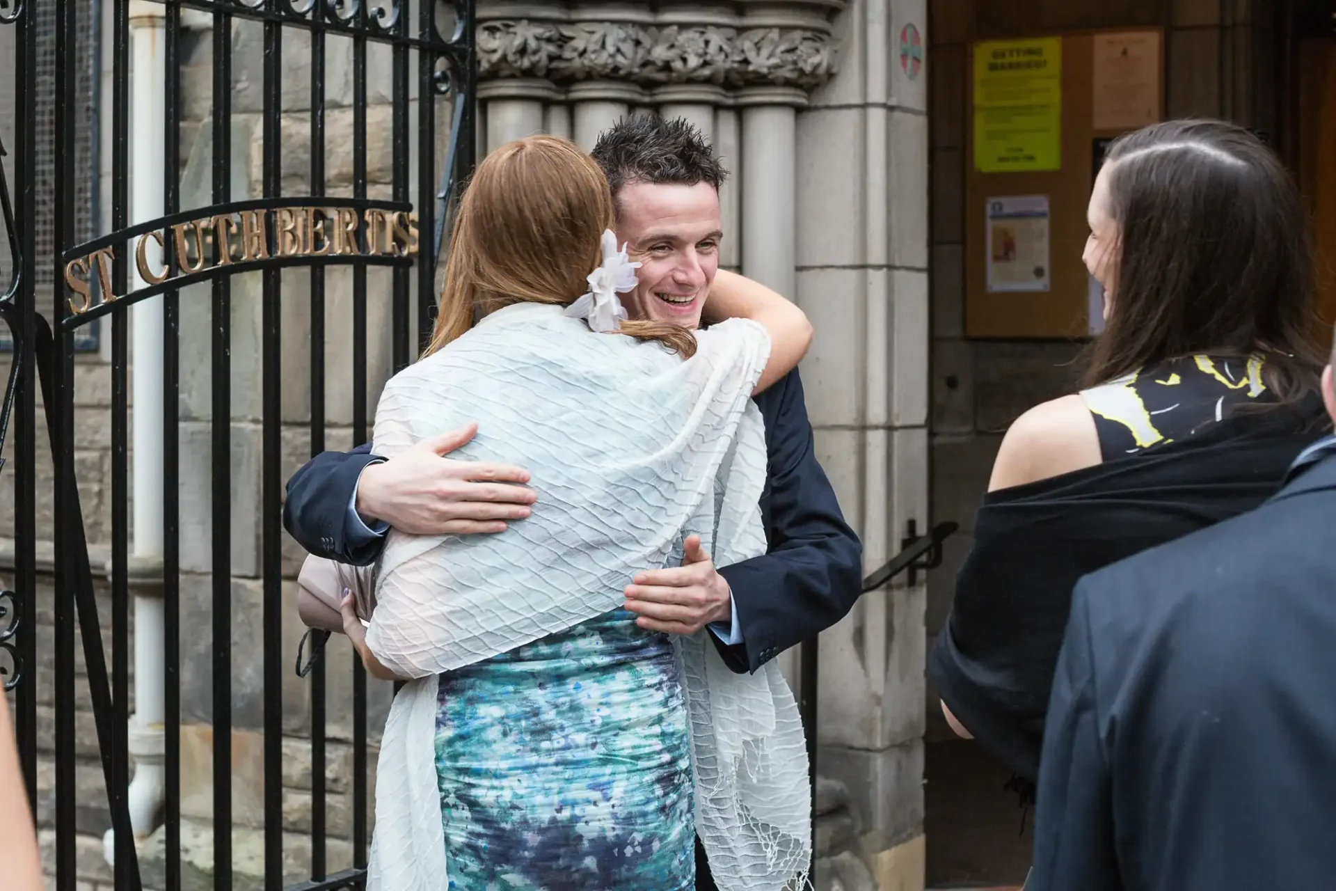 A man and a woman embracing joyfully outside a building, while another woman watches them.