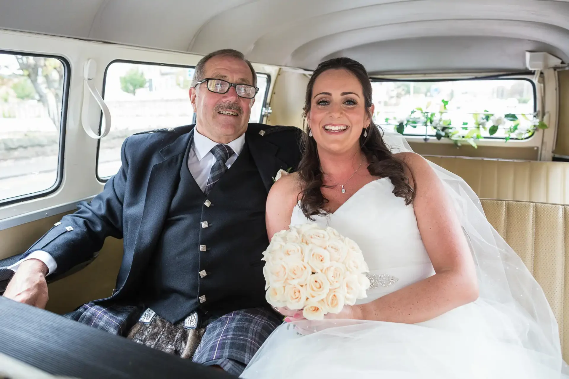 A bride in a white dress holding a bouquet sits beside an older man in a kilt inside a vintage car.