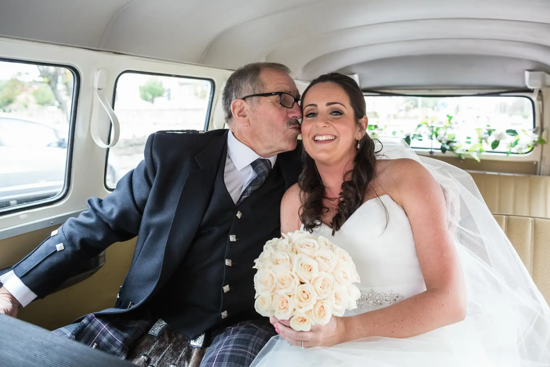 A bride in a white dress holding a bouquet smiles as she receives a kiss on the cheek from an older man in a suit and kilt inside a vintage car.