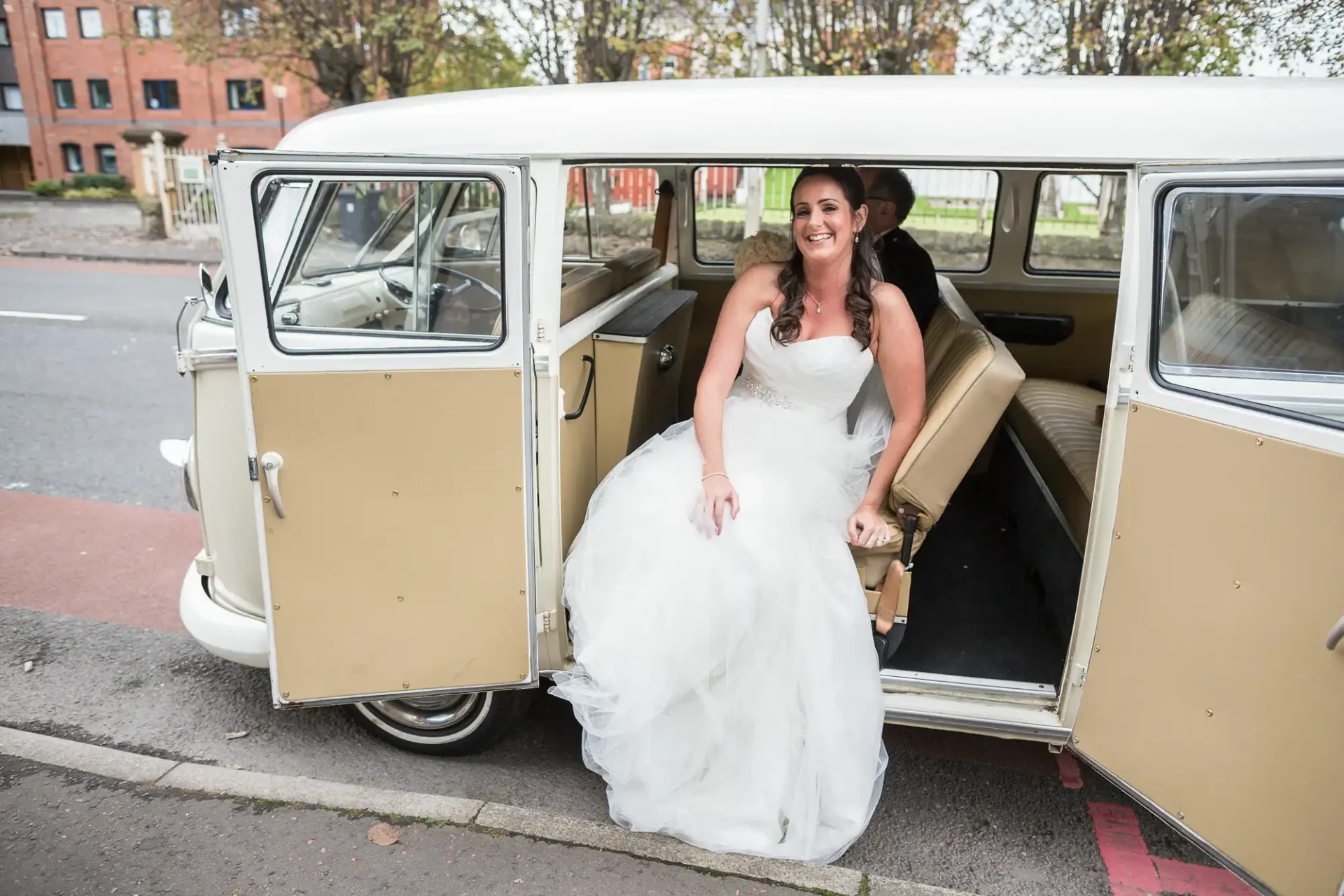 Bride in a white dress smiling as she exits a vintage van on a city street.