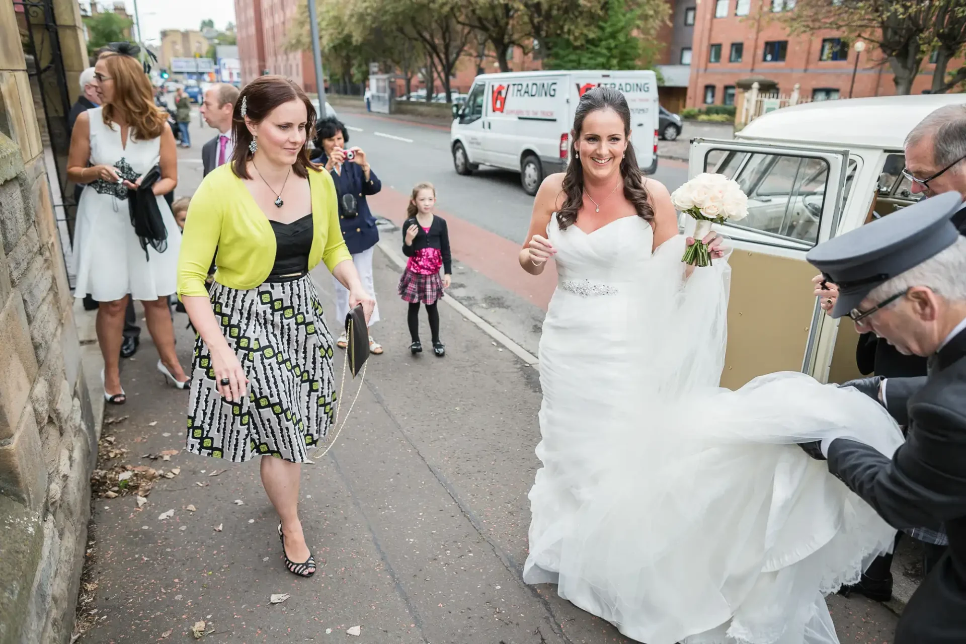A bride in a white gown exits a vintage car on a city street, assisted by an older man, as a woman in a patterned dress and yellow cardigan approaches smiling.