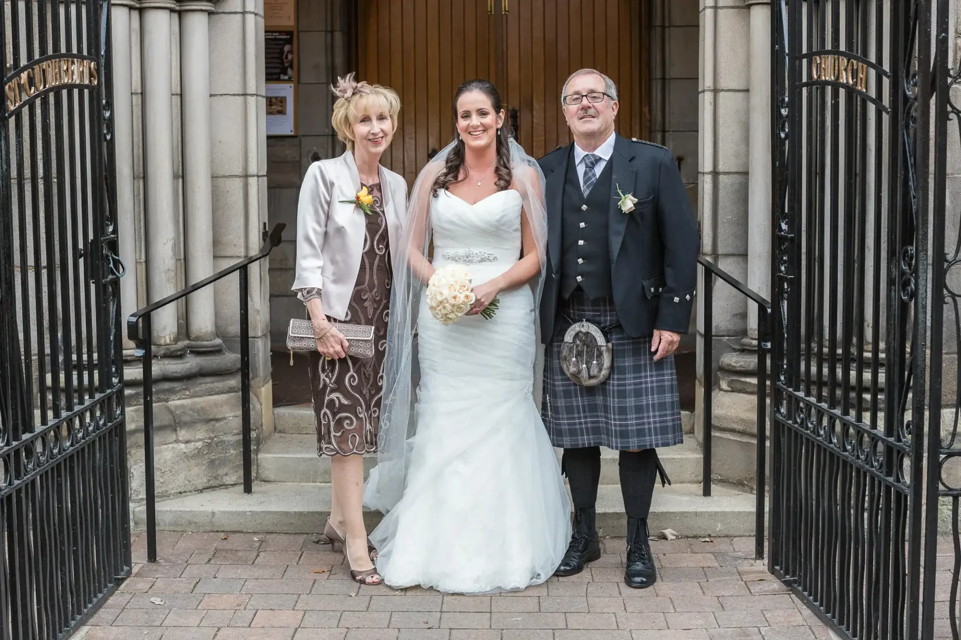 A bride in a white dress stands with an older couple dressed in formal attire, including tartan elements, outside a church gate.