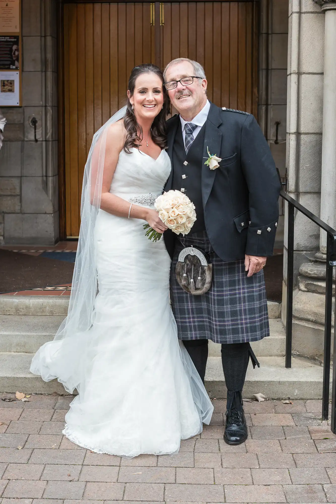 Bride in a white dress and groom in a kilt smiling outside a church entrance.