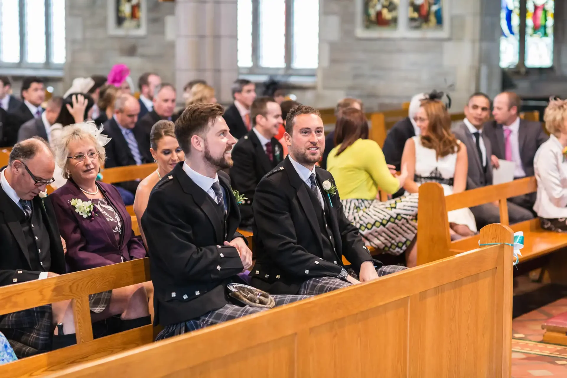 Two men wearing traditional kilts sitting in a church pew during a wedding ceremony, surrounded by other guests.