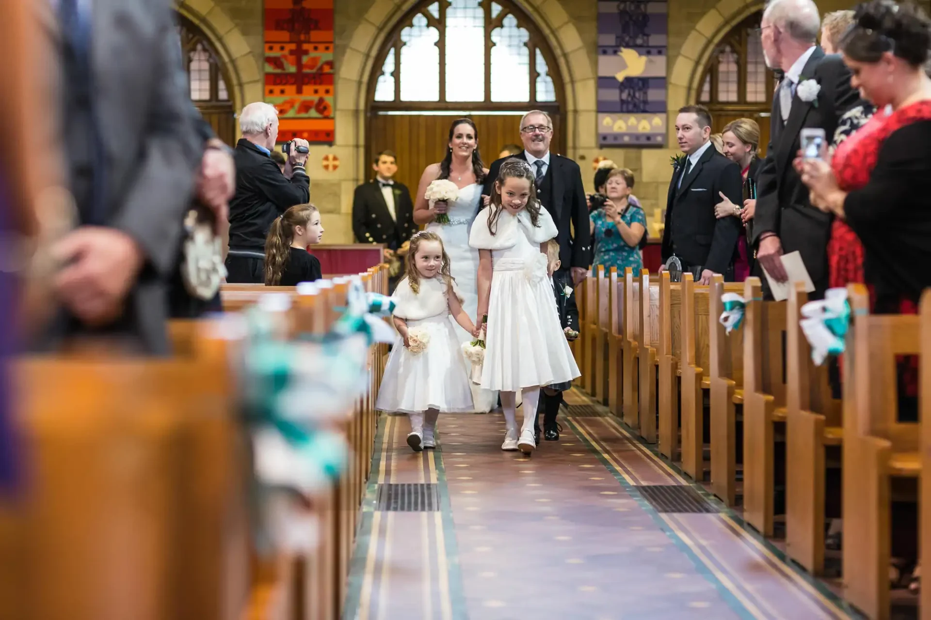 A joyful procession down a church aisle with a man and two girls in white dresses, surrounded by elegantly dressed guests.