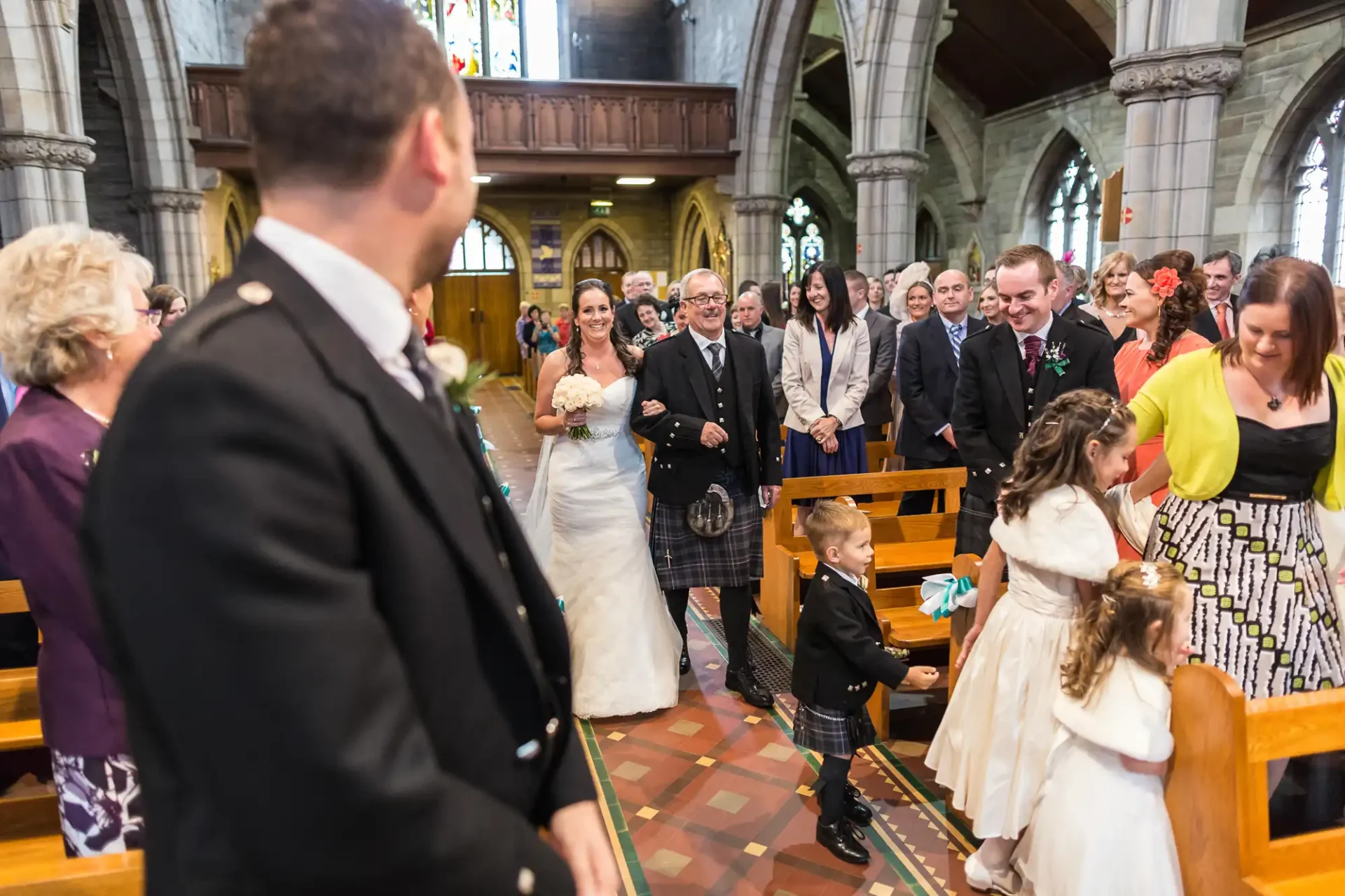 Bride walking down the aisle with her father in a church, as guests watch and a groom awaits at the altar, some wearing traditional scottish attire.