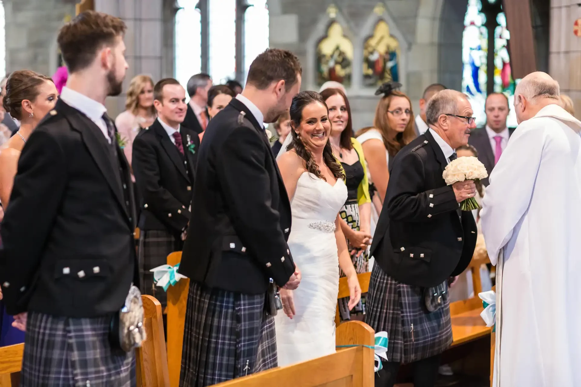 Bride and groom holding hands during a wedding ceremony in a church, surrounded by guests in traditional scottish attire.