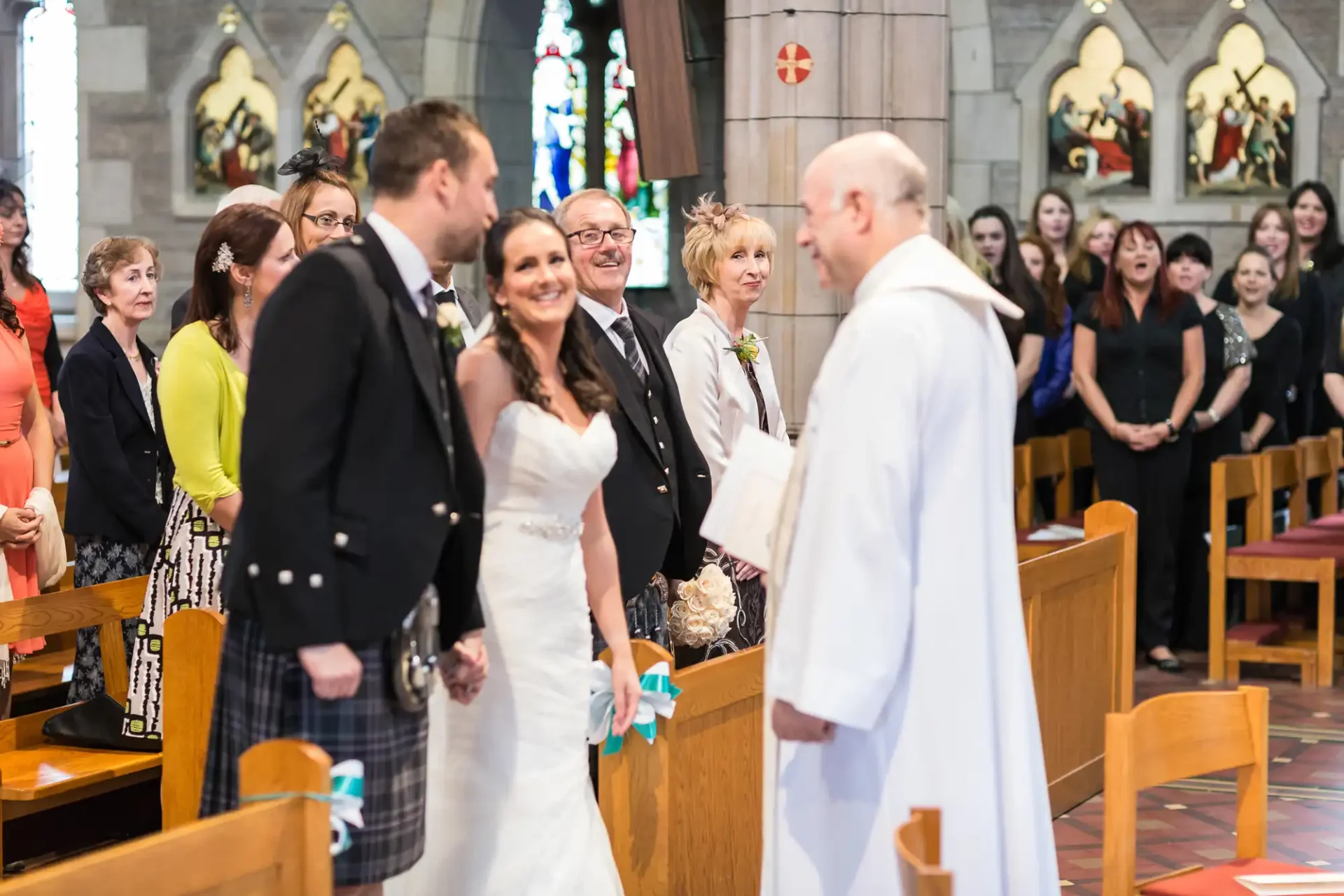 A bride and groom smiling at each other in a church during their wedding ceremony, surrounded by guests and a priest.