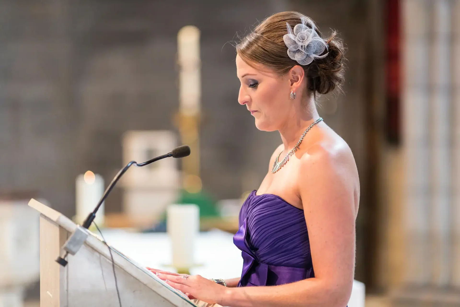 A woman in a purple dress with an ornamental flower in her hair speaks at a podium in a softly lit hall.