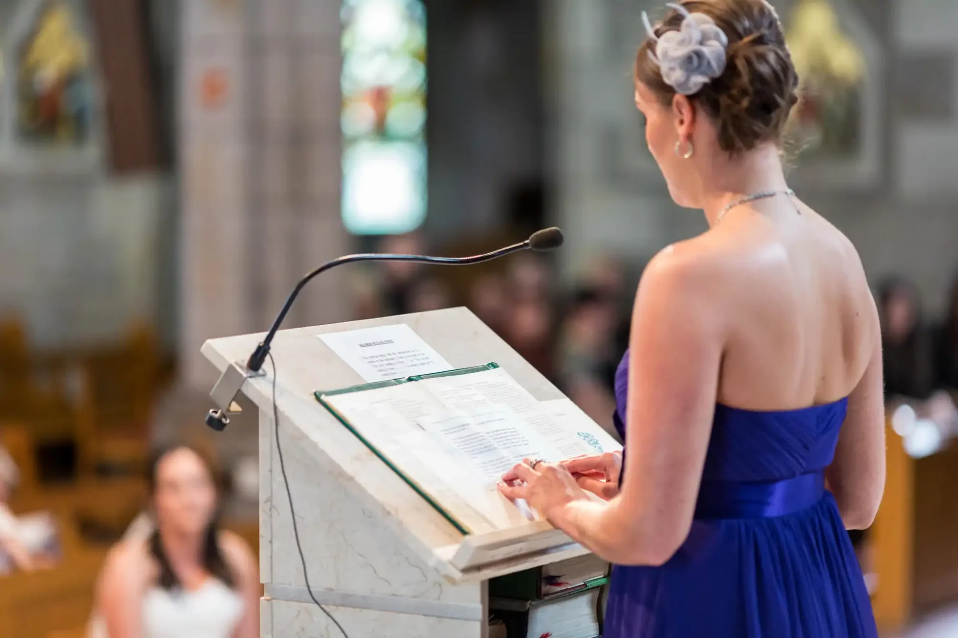 Woman in blue dress standing at lectern in church, reading from a book during a ceremony, with blurred background.