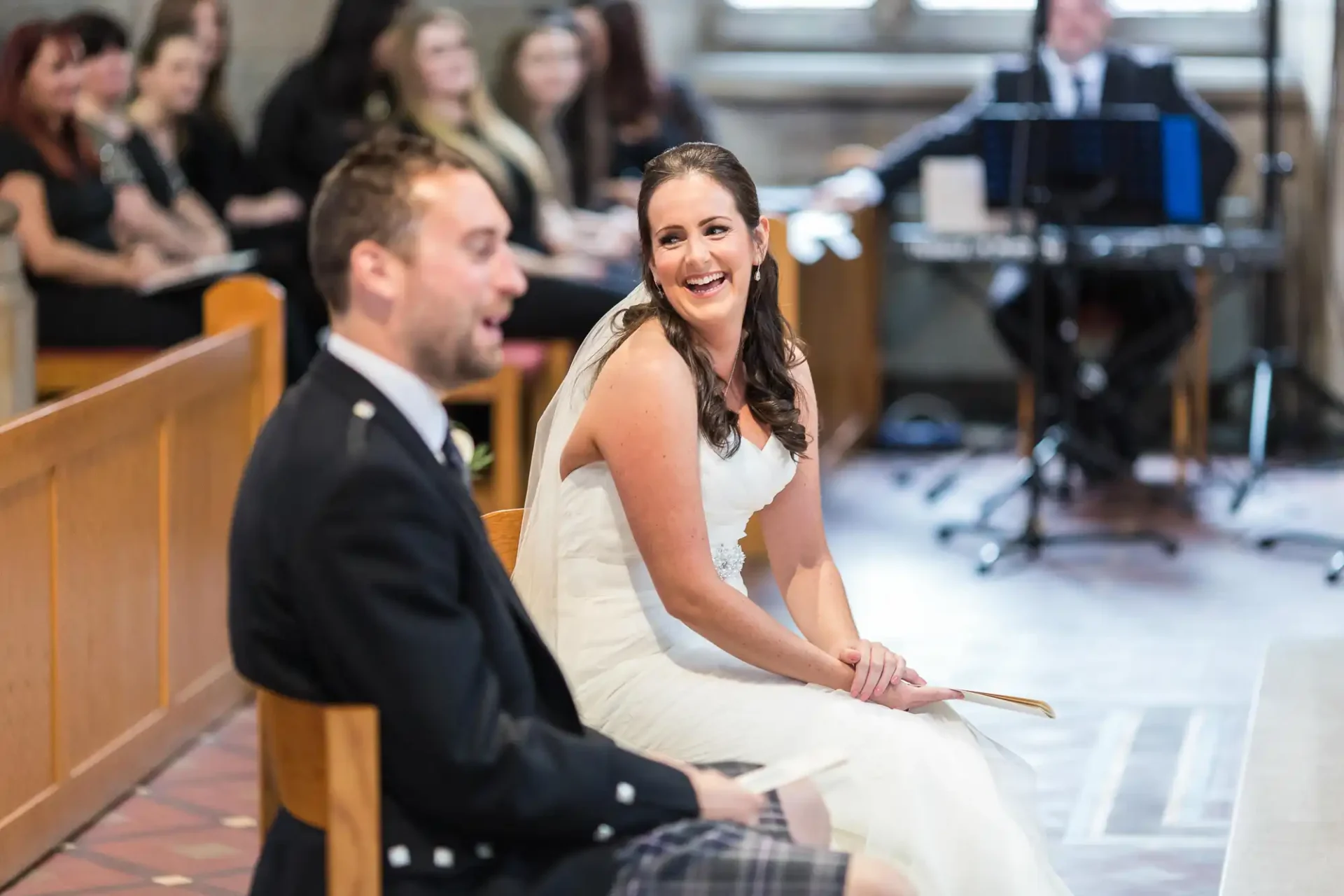 A bride and groom laughing joyfully in a church, seated during their wedding ceremony, with guests looking on.