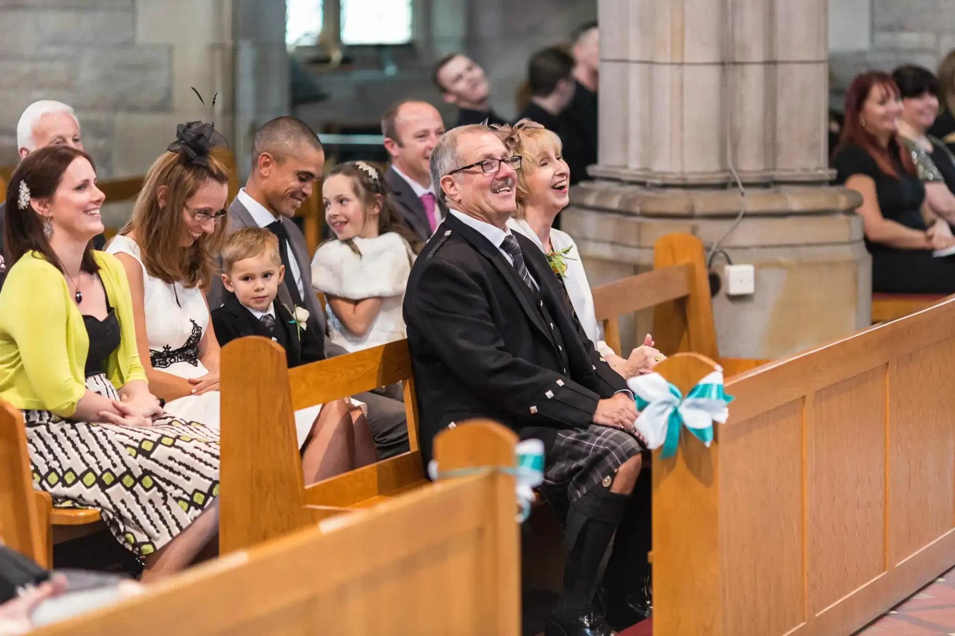 Guests in formal attire smiling and watching an event inside a church, including a man in a kilt and a woman with a corsage.