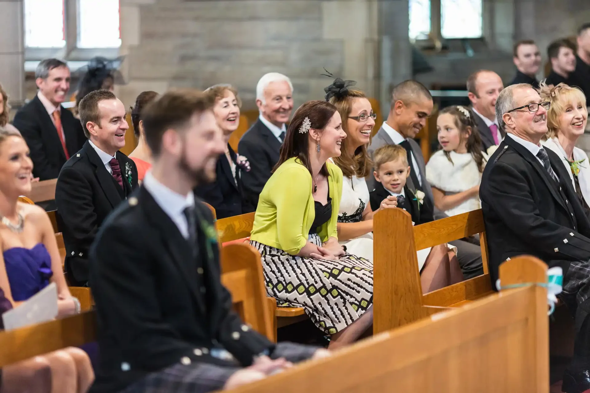 Guests in formal attire smiling and seated in church pews during a wedding ceremony.