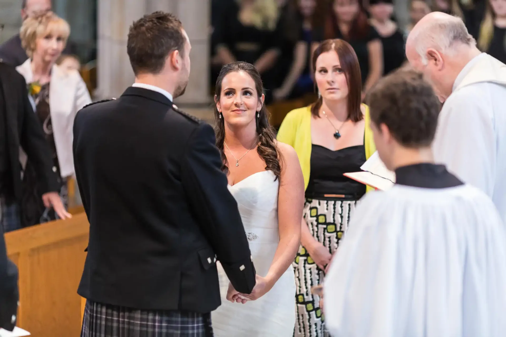 A bride and groom holding hands at the altar, facing each other, with a priest and guests in the background.