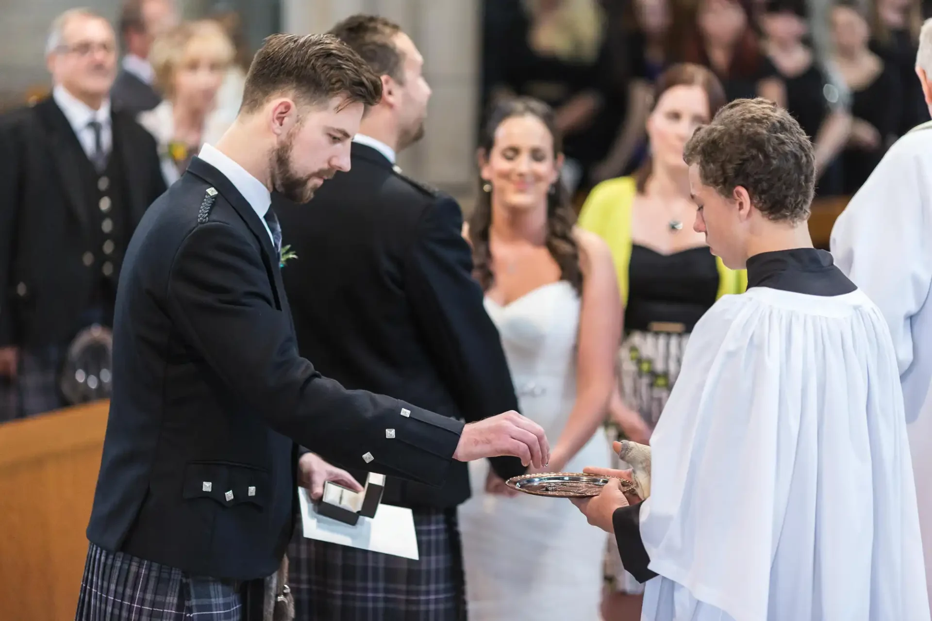A groom in a kilt exchanging rings with a bride during a church wedding ceremony, witnessed by guests and a clergy member.