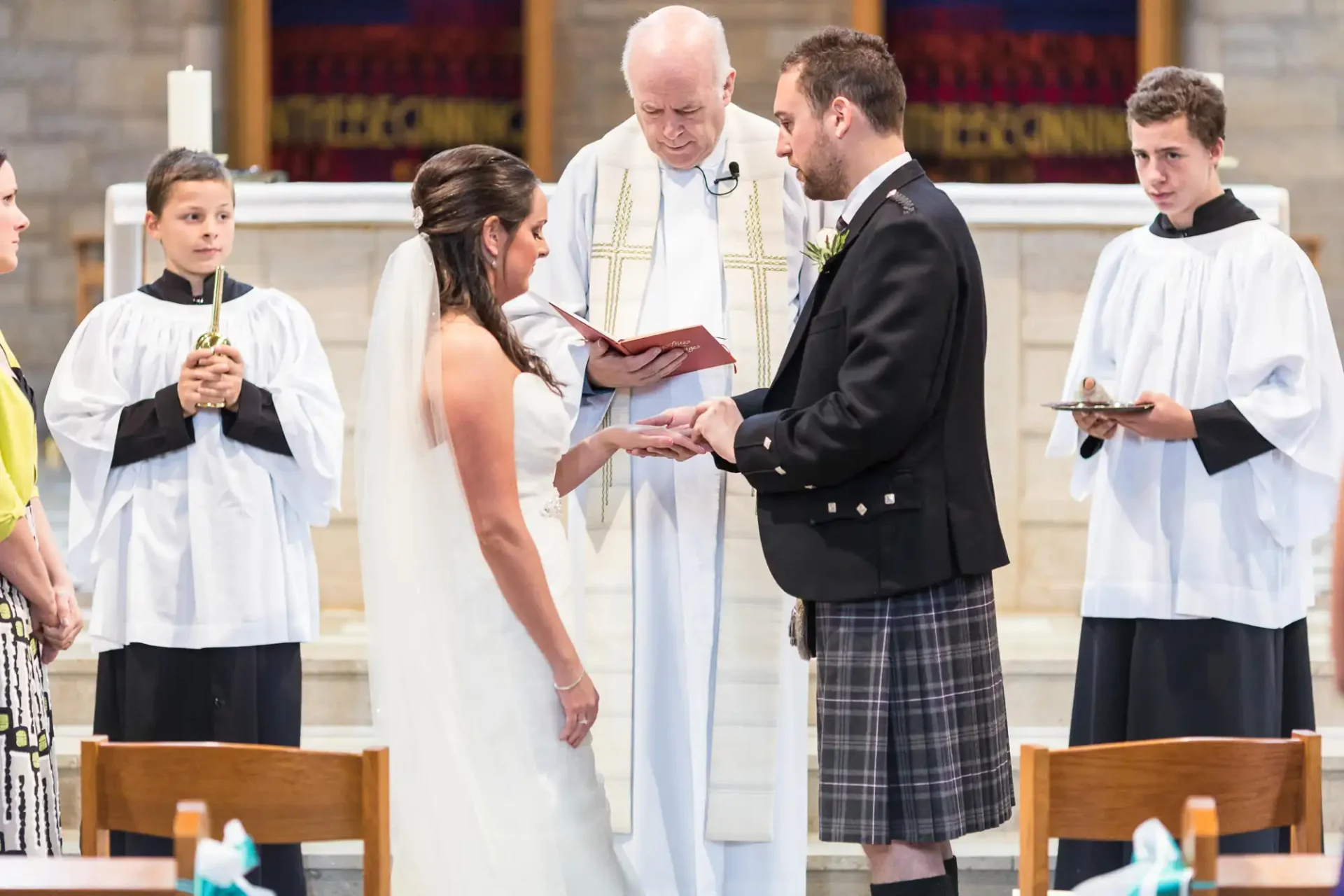 A bride and groom exchange vows in a church ceremony, officiated by a priest, with two altar boys watching.