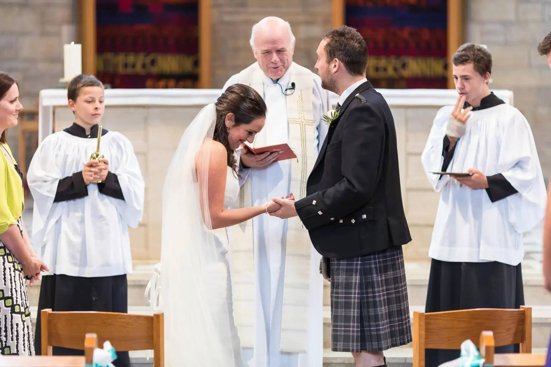 Bride and groom exchanging rings at a church wedding ceremony, officiated by a priest, with attendants watching in the background.