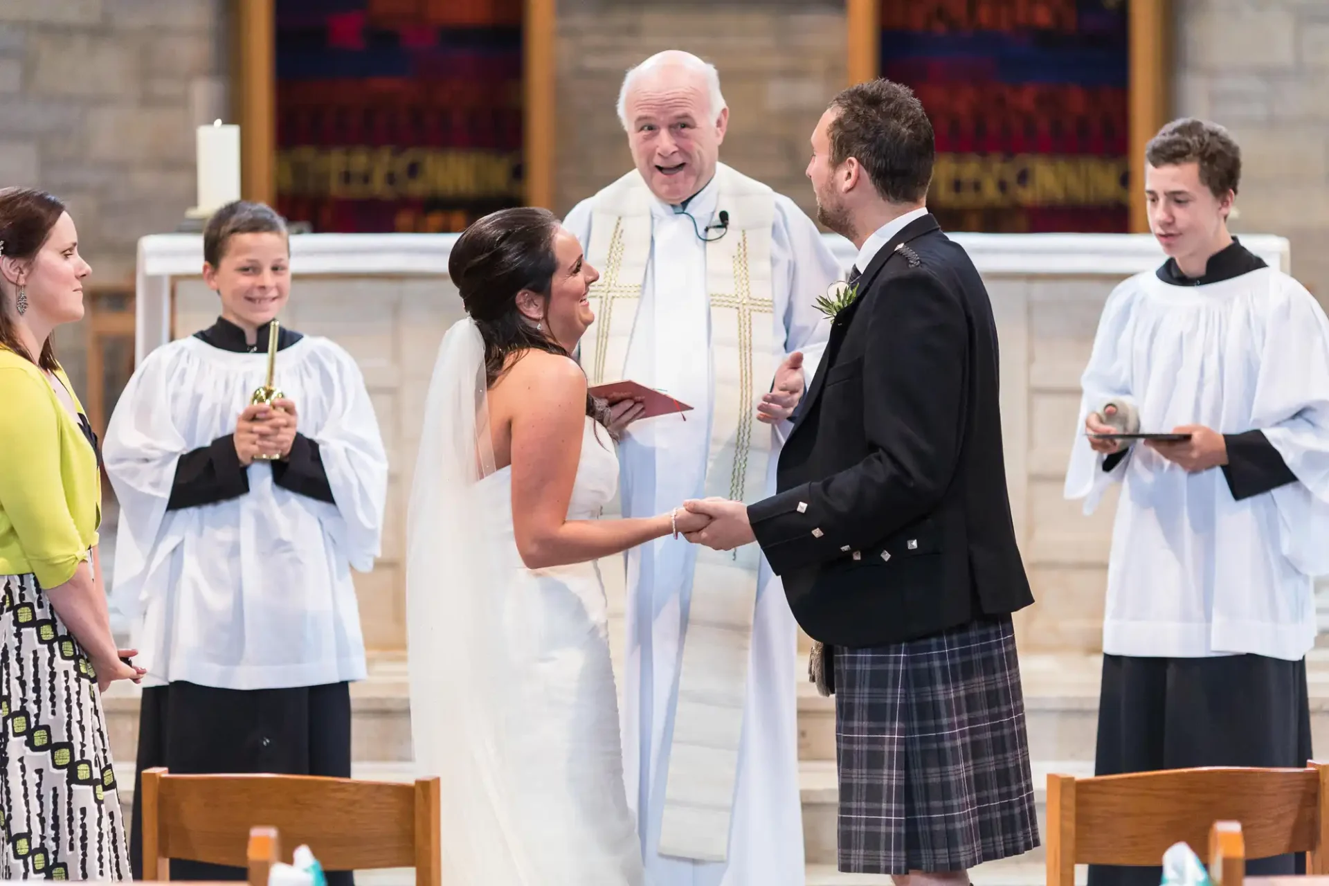 A bride and groom exchange vows in a church, officiated by a priest, with two attendants in white robes to the side.