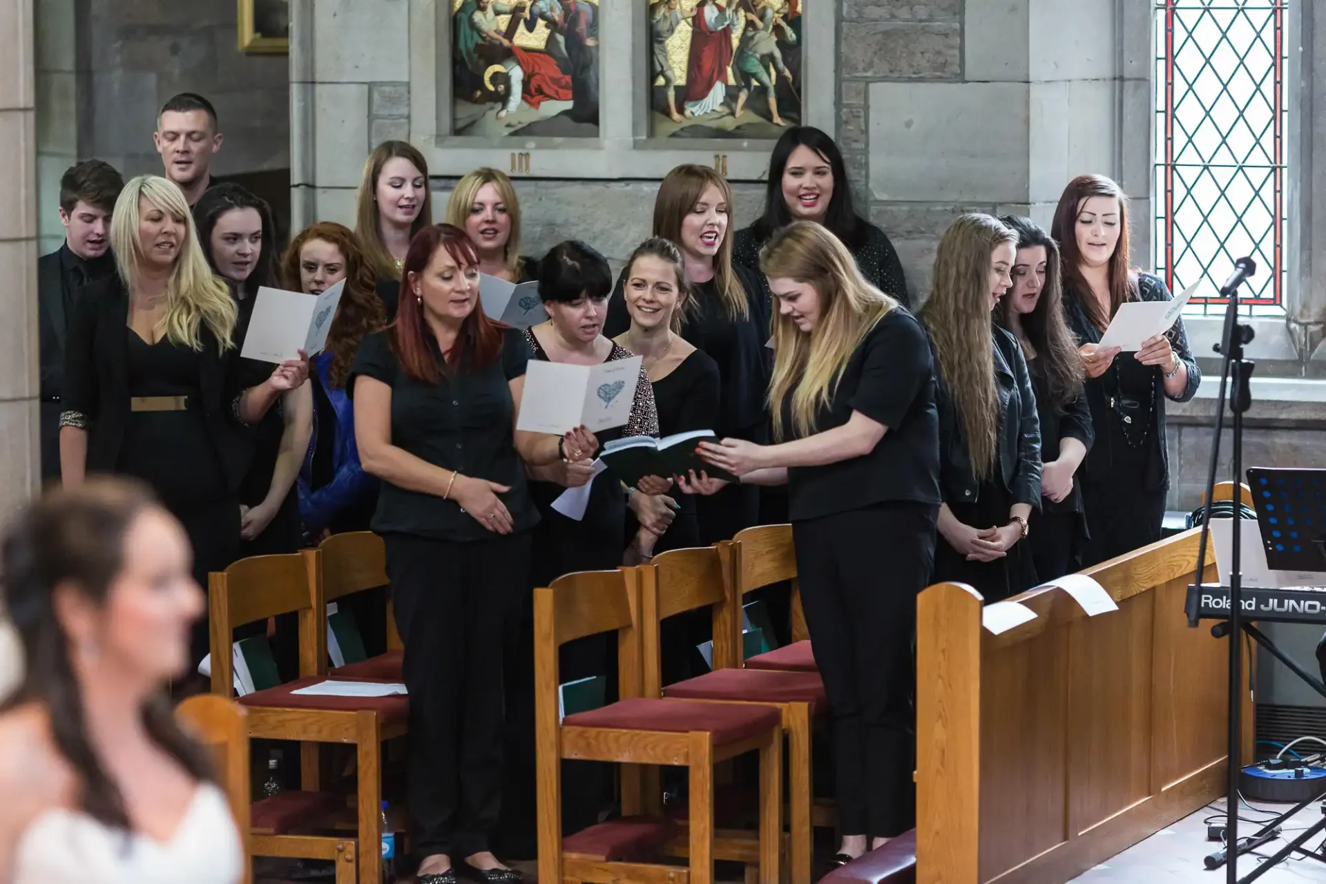 A choir of mixed young adults sings from sheet music inside a church, with attentive audience members seated nearby.