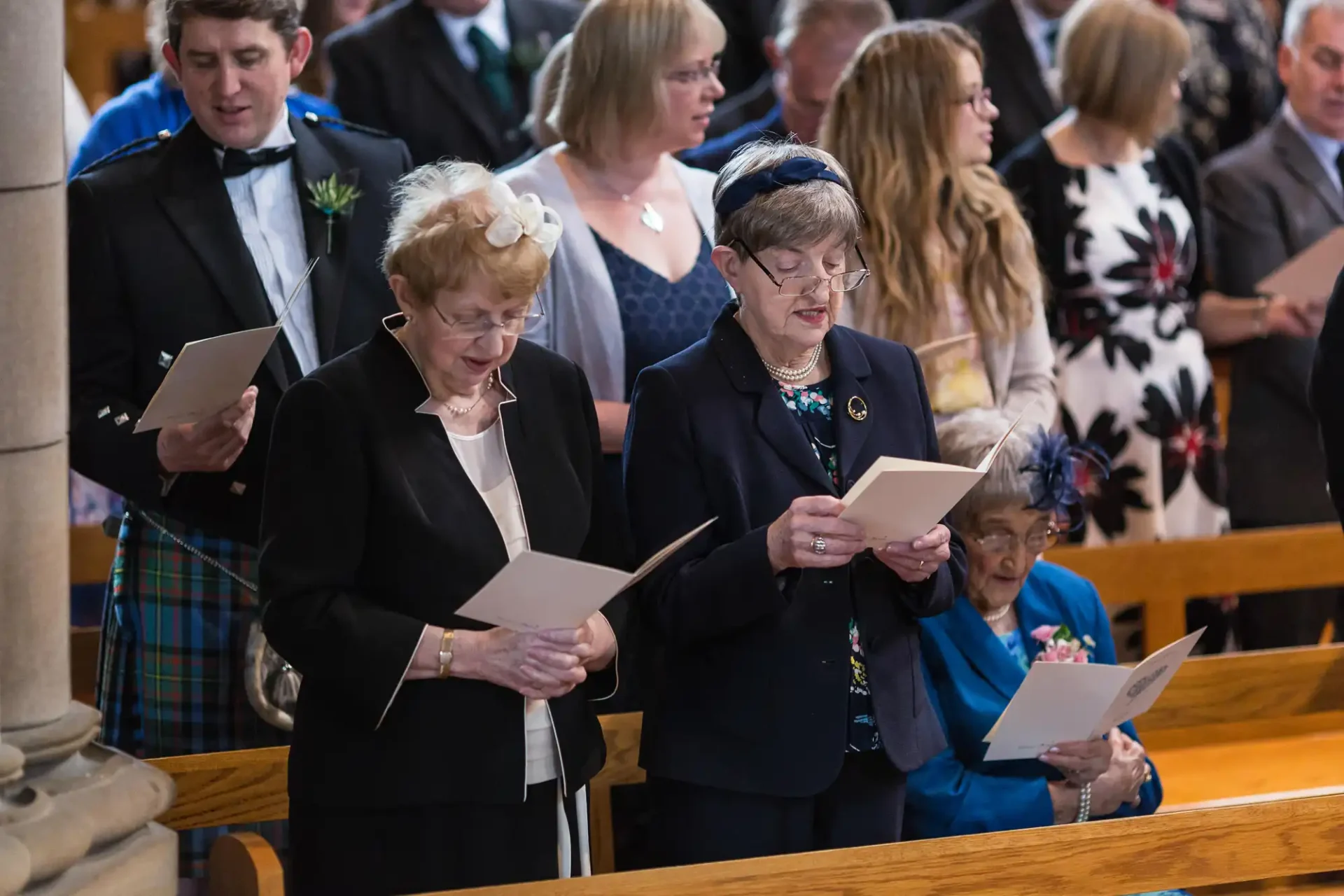 Two elderly women reading from hymnals during a church service, surrounded by other attendees in formal attire.