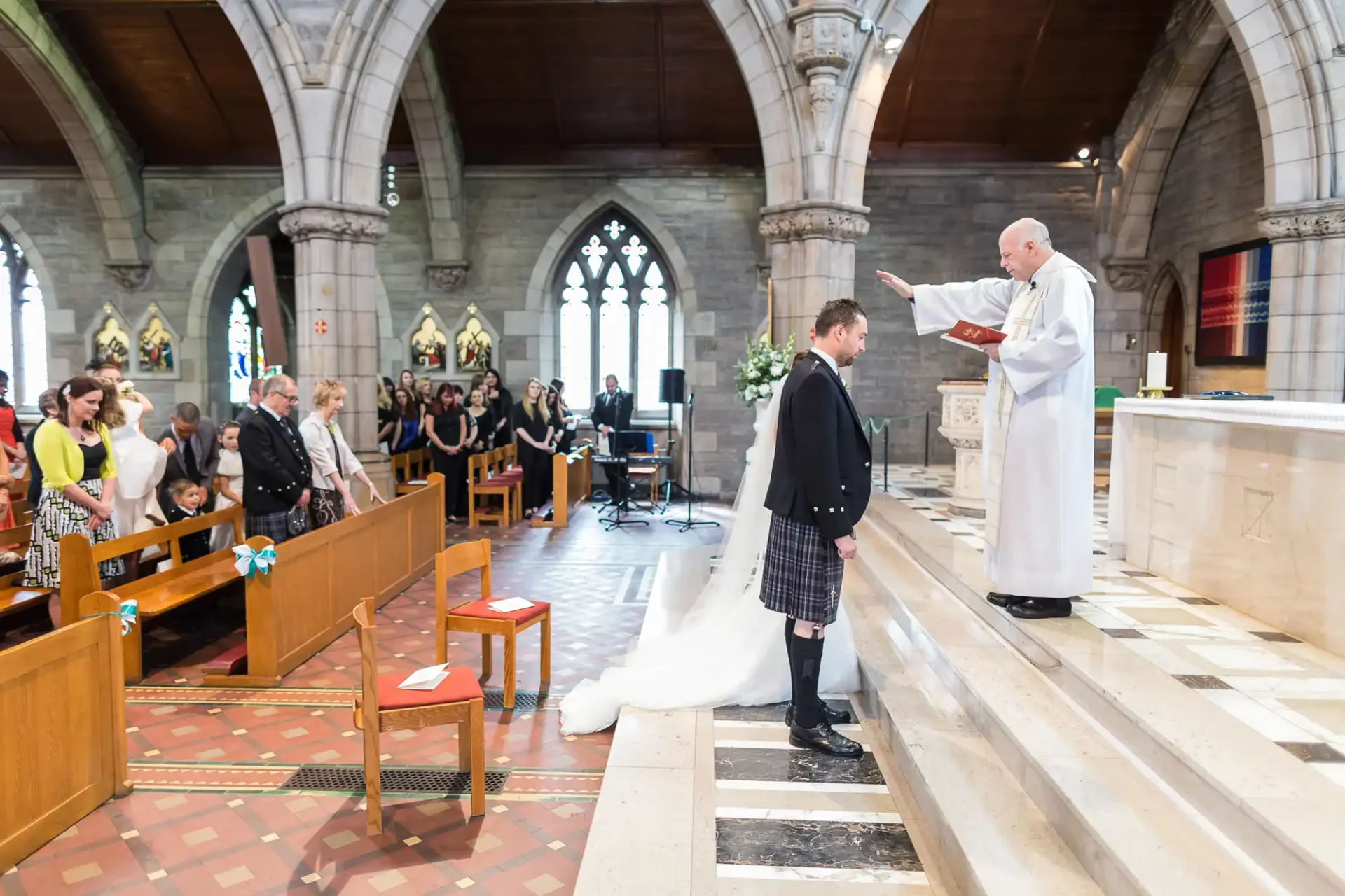 A wedding ceremony in a church with a priest speaking to a groom in a kilt, surrounded by guests seated in pews.