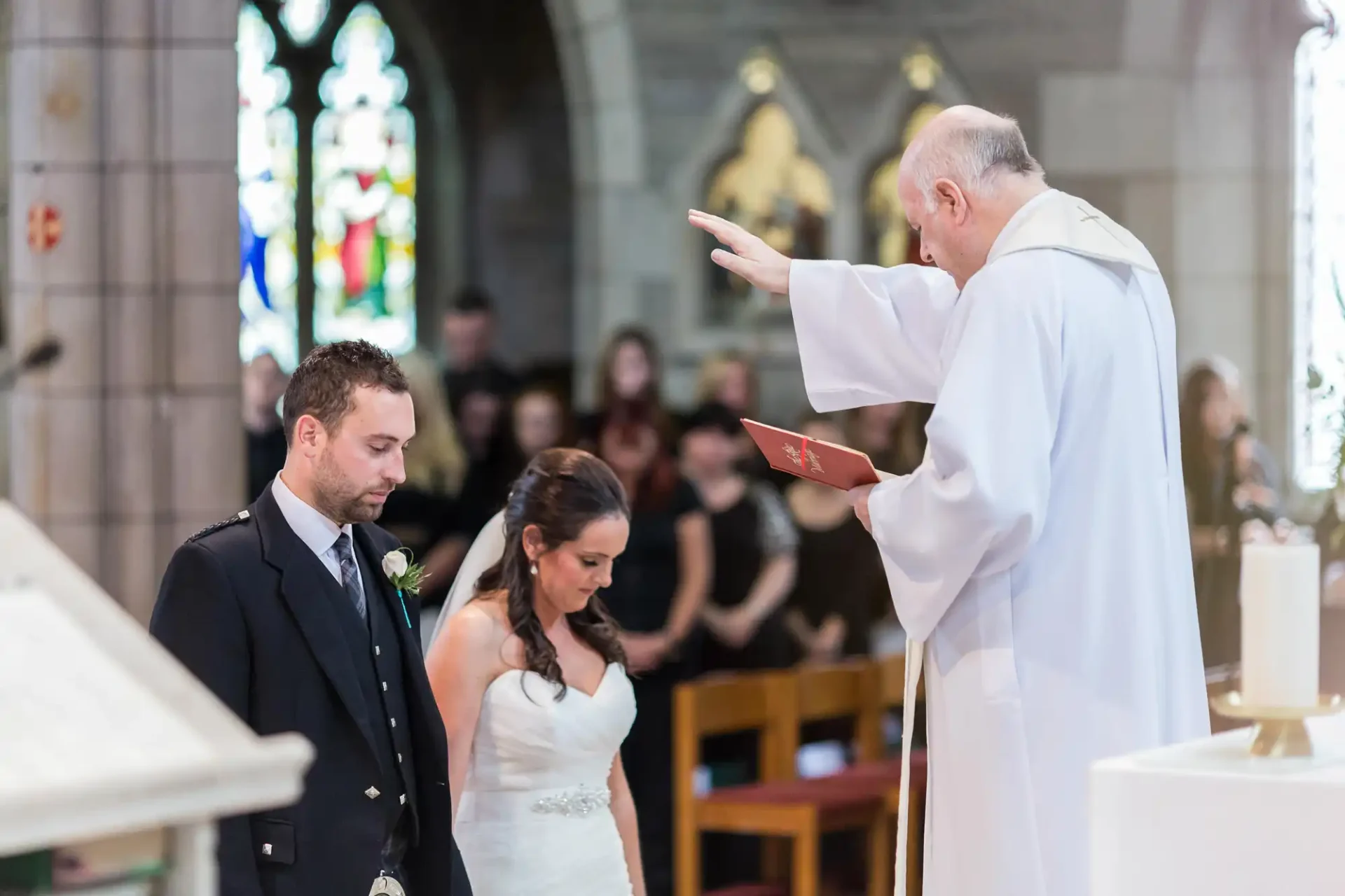 A priest conducting a wedding ceremony blesses a couple standing at the altar in a church, with guests in the background.