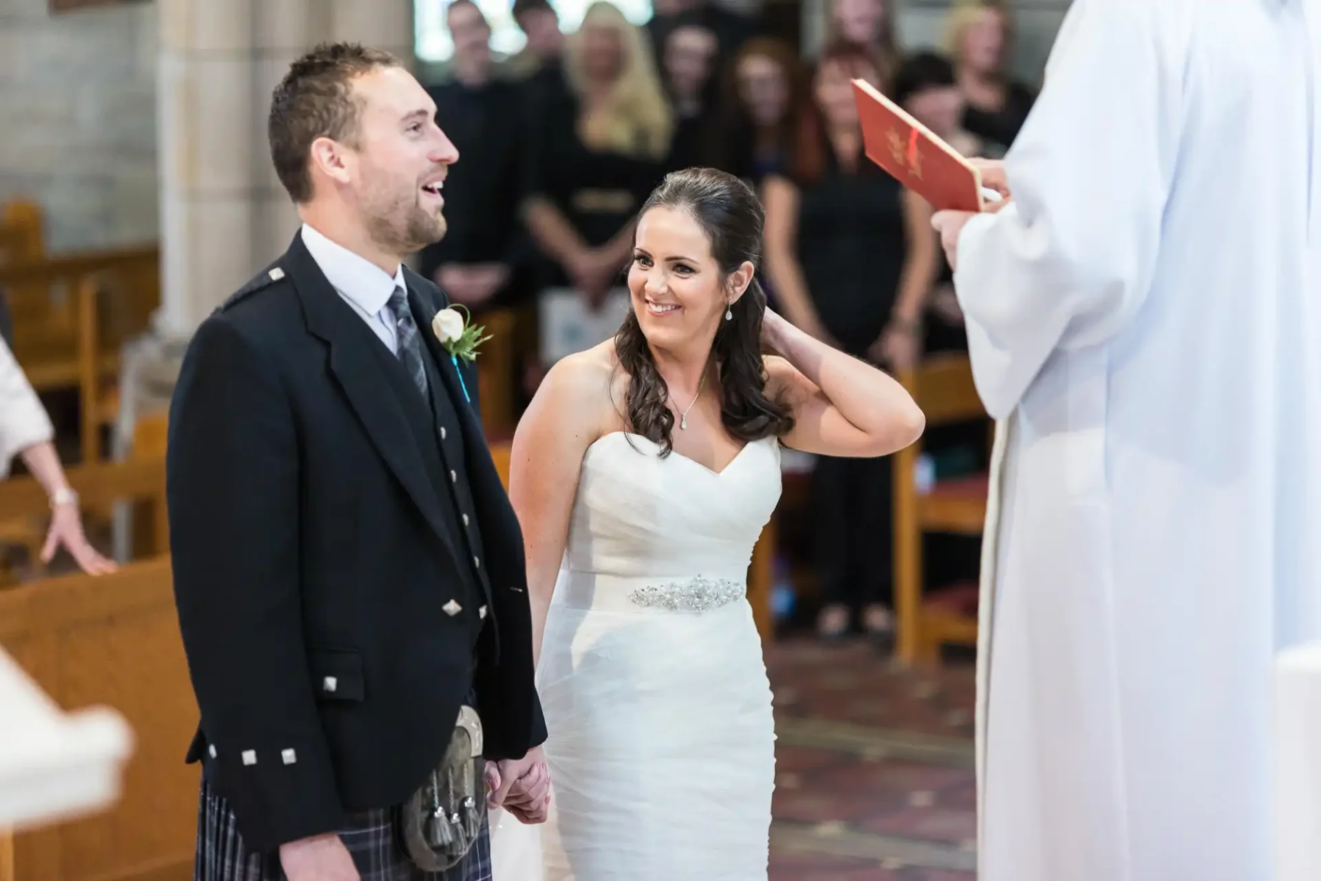Bride and groom smiling at each other during a wedding ceremony in a church, with a priest leading the ceremony in the foreground.