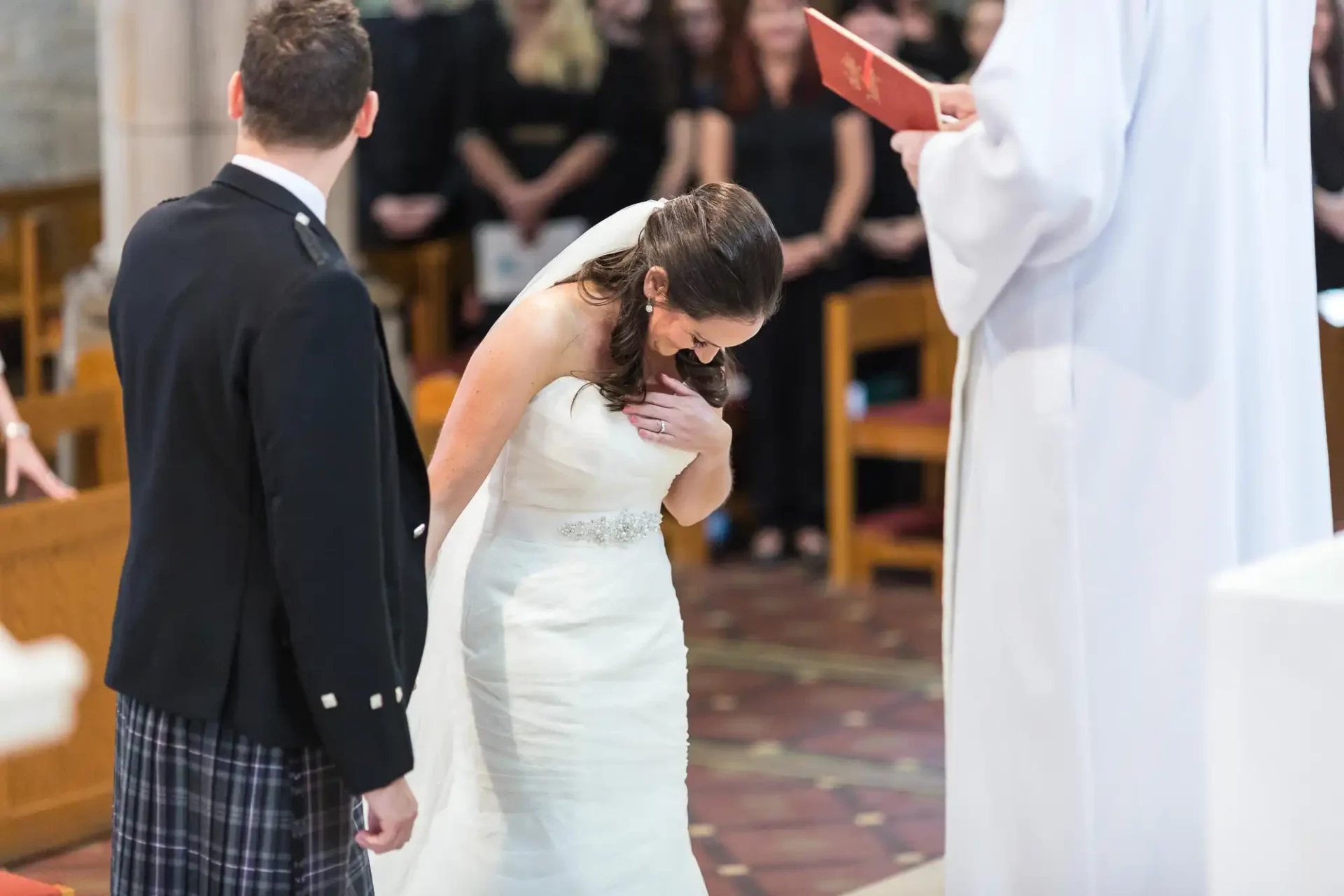 Bride in a white dress bowing her head during a church wedding ceremony, standing next to a groom in a kilt, with a priest and attendees in the background.