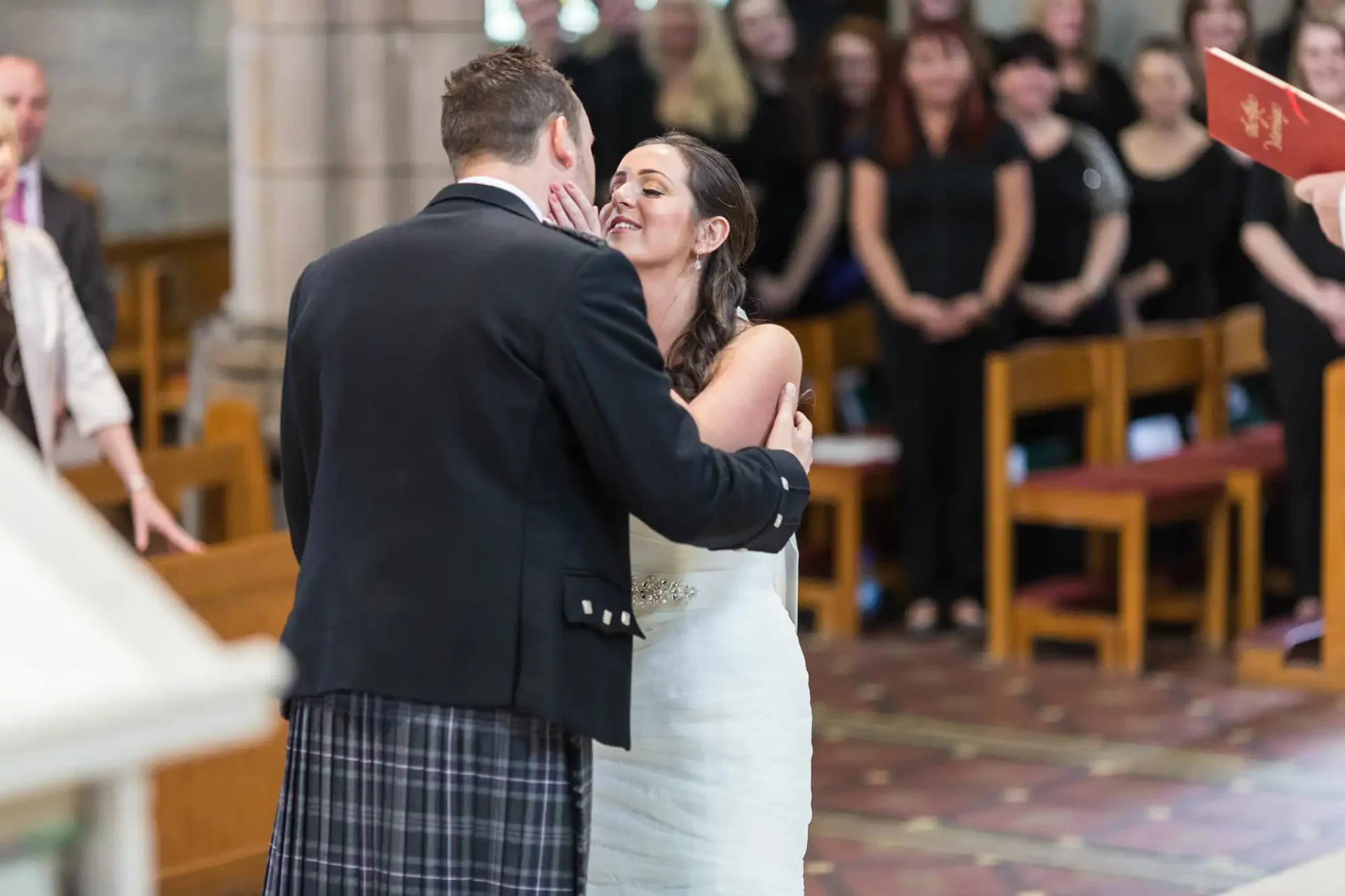 A bride and groom, the groom in a kilt, embrace during their wedding ceremony inside a church, with guests looking on.