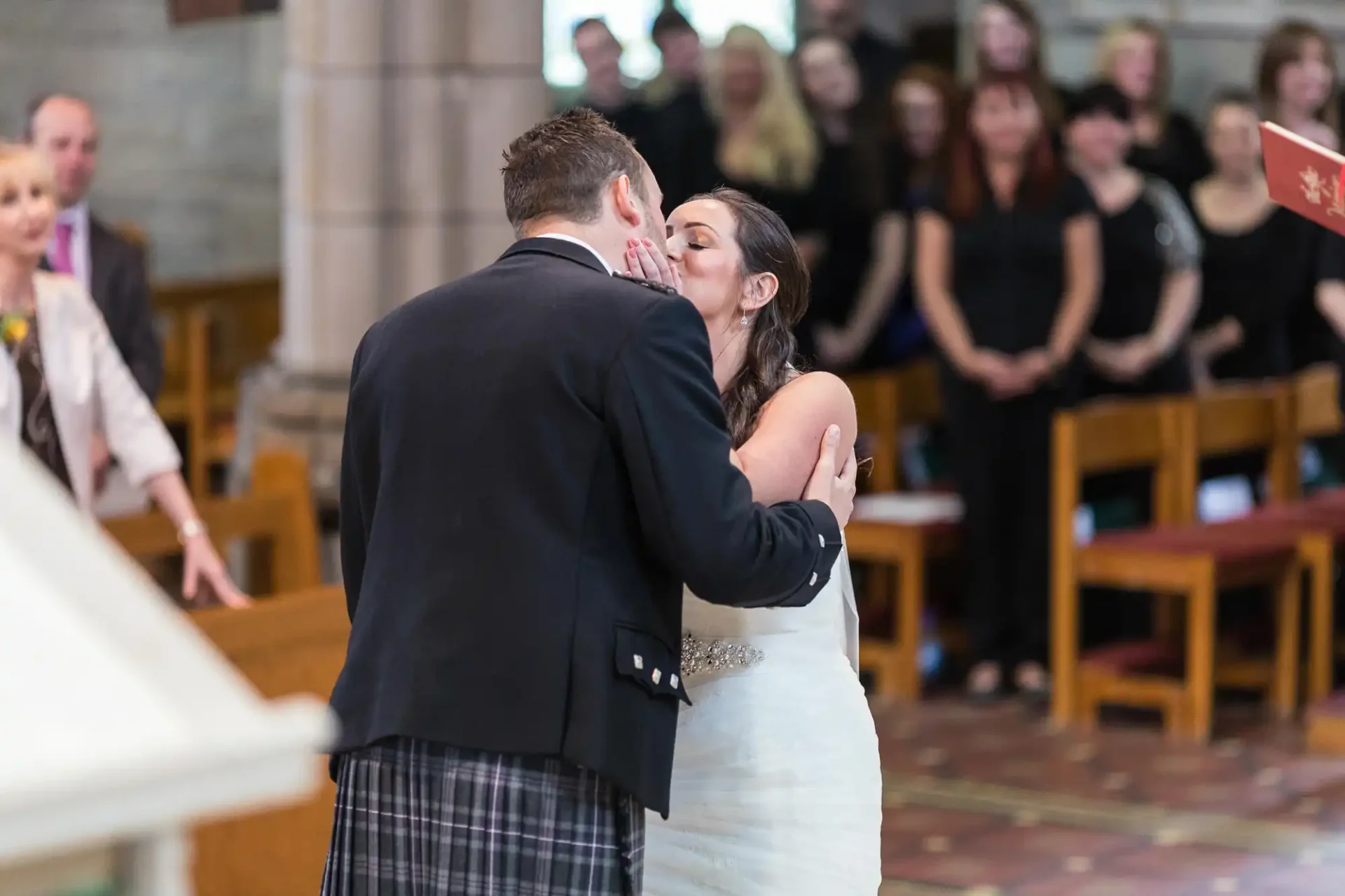 A bride and groom kissing at the altar in a church, with guests in the background and a man in a kilt visible.