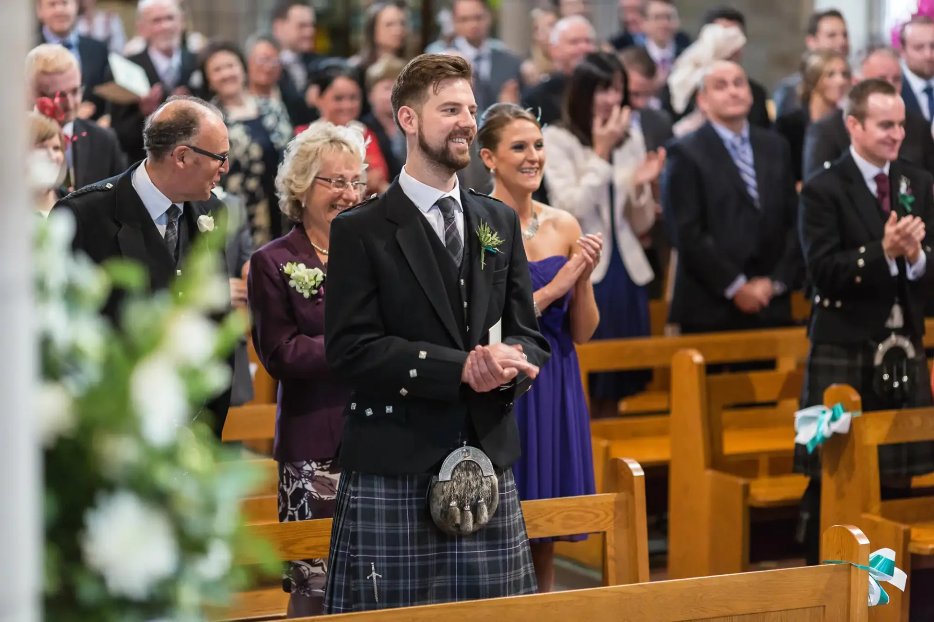 A groom in a kilt smiles during a wedding ceremony inside a church, surrounded by guests in formal attire.