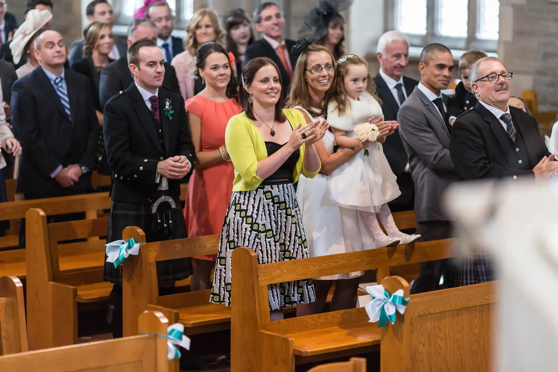 Guests smiling and clapping at a wedding ceremony inside a church with wooden pews decorated with bows.
