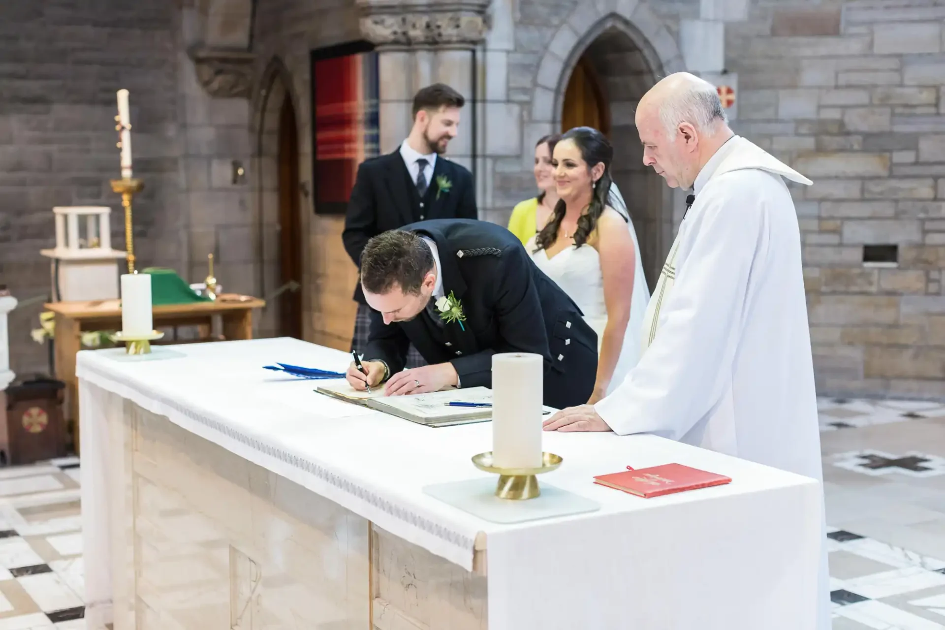 Groom signing the marriage register in a church, observed by a priest and bride, alongside a witness in the background.