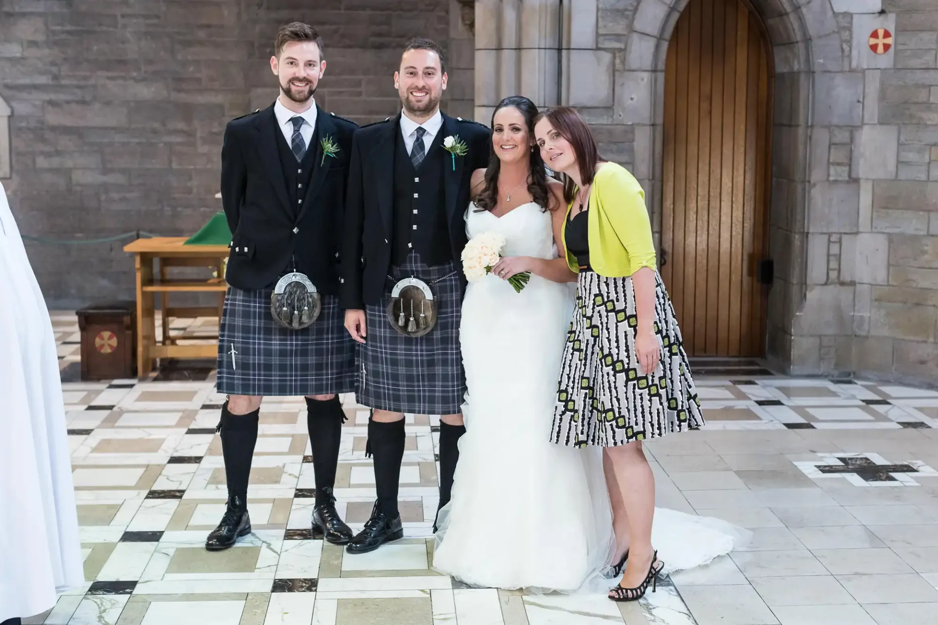 Two men in kilts and a bride and bridesmaid pose smiling inside a church.