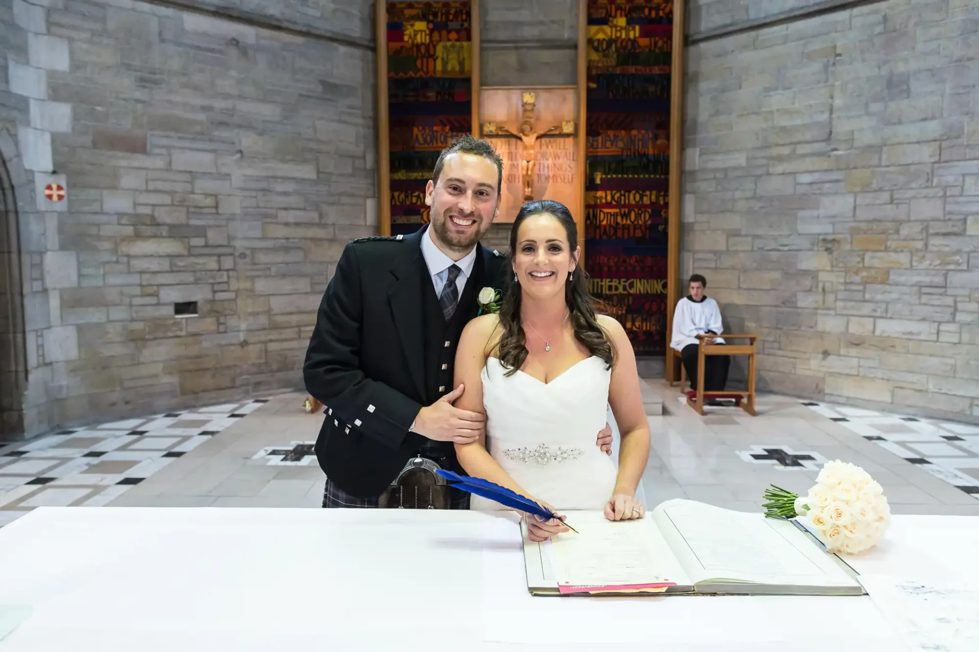A bride and groom smiling at the camera while signing a registry book in a church with stained glass windows in the background.
