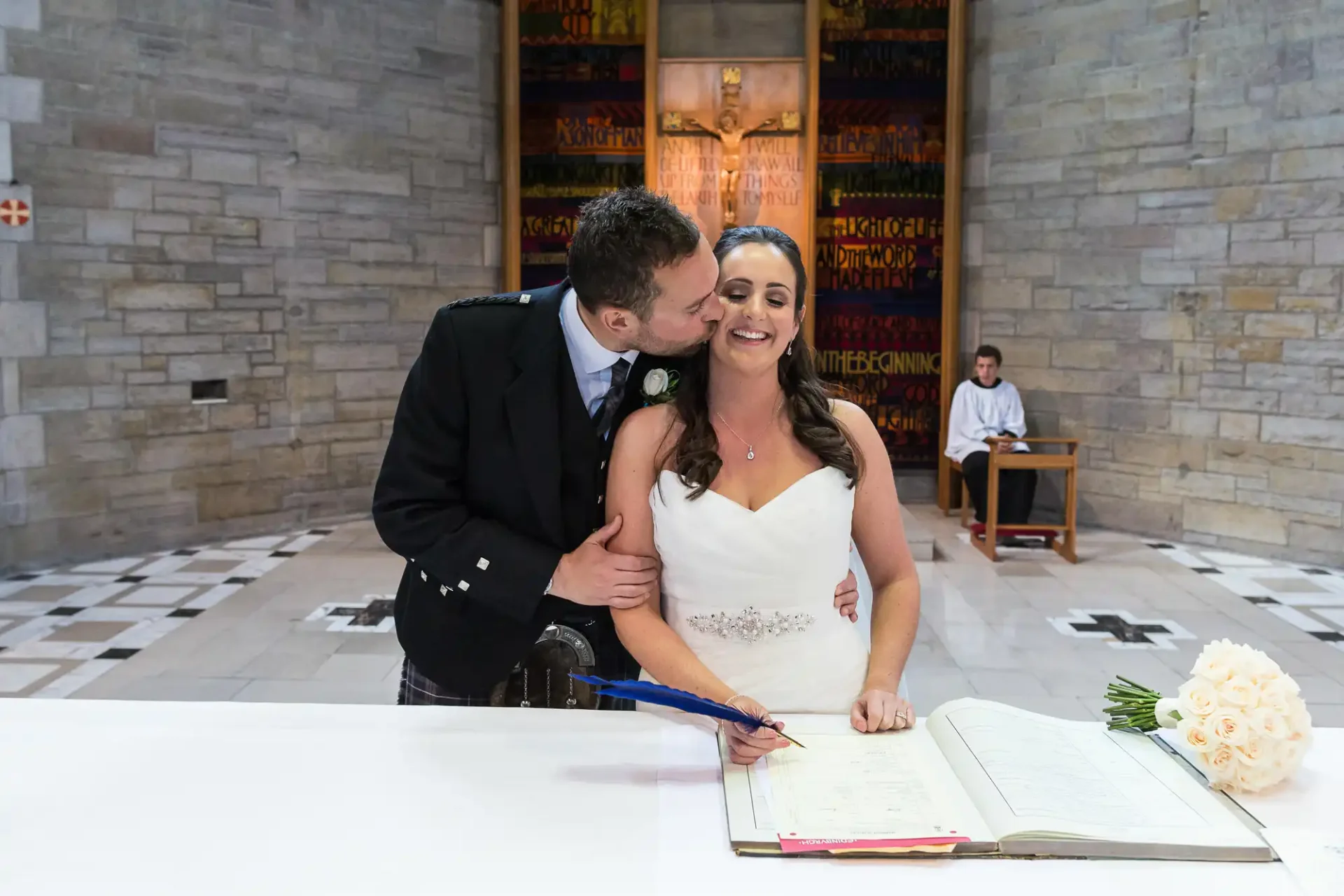 Groom kissing bride on the cheek as she signs a document at a table in a church, with a pianist in the background.