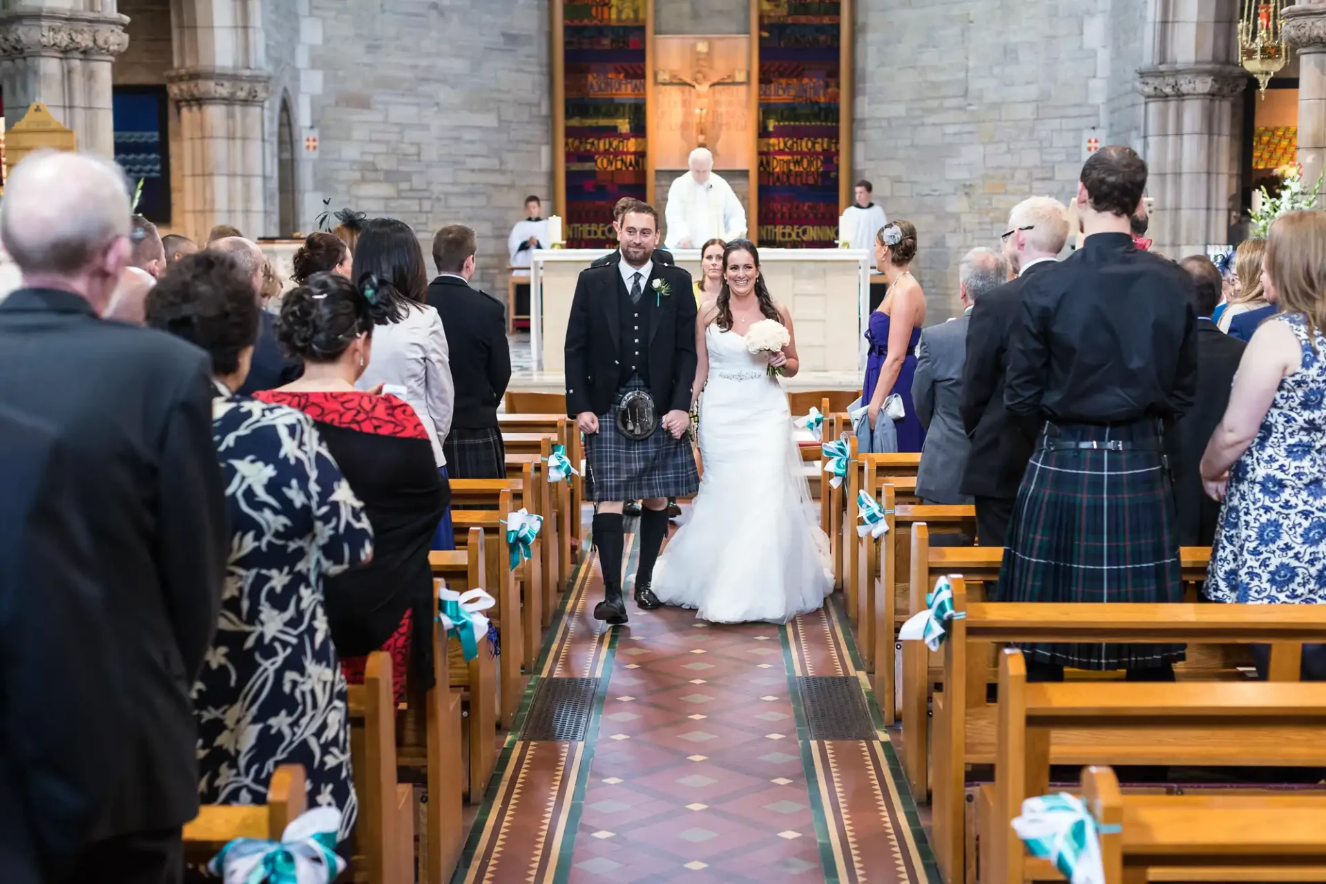 A bride and groom walk down the aisle of a church, guests watching, groom in a kilt, blue ribbon decorations on pews.