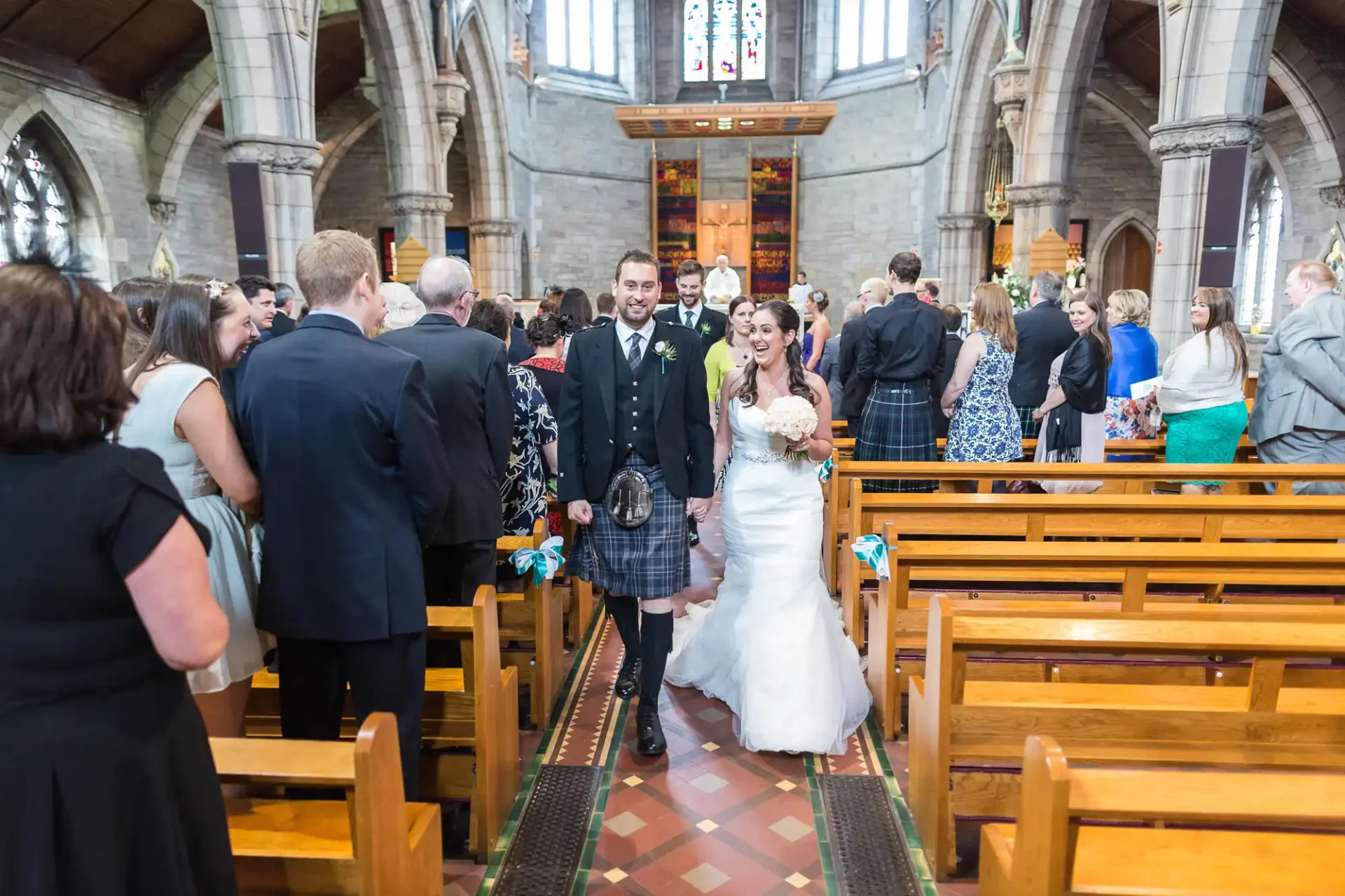 Newlywed couple smiling as they walk down the aisle in a church, surrounded by guests, with the groom in a kilt.