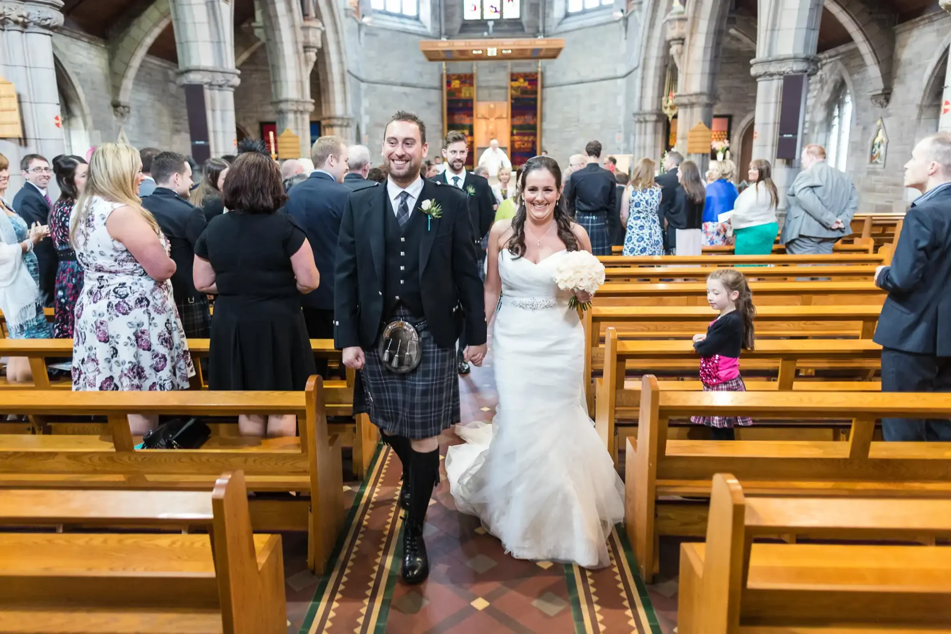 A newly married couple smiling as they walk down the aisle of a church, surrounded by guests. the groom wears a kilt and the bride wears a traditional white gown.