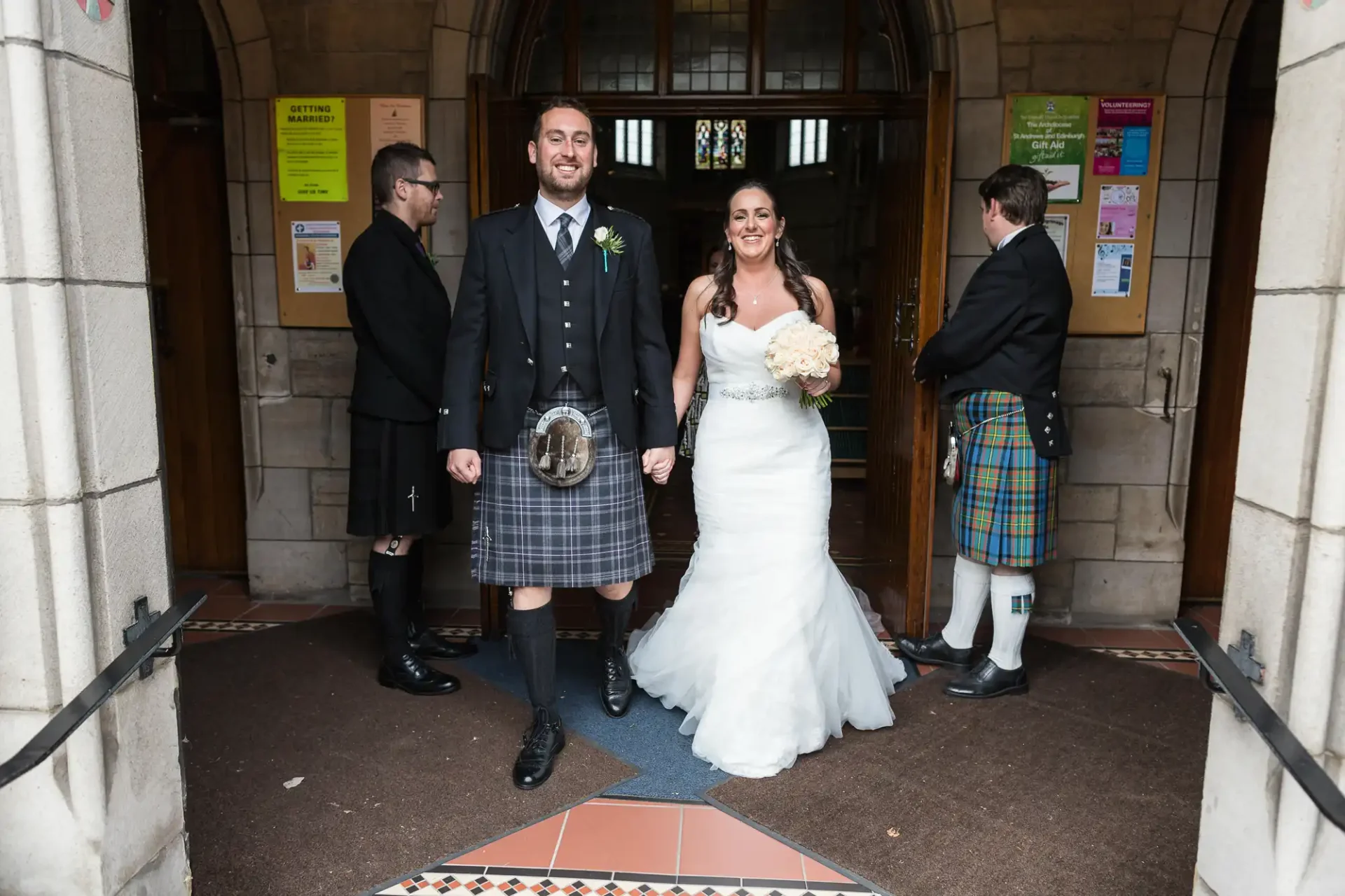 A smiling bride and groom, dressed traditionally (groom in a kilt), exit a church, flanked by two ushers in kilts.