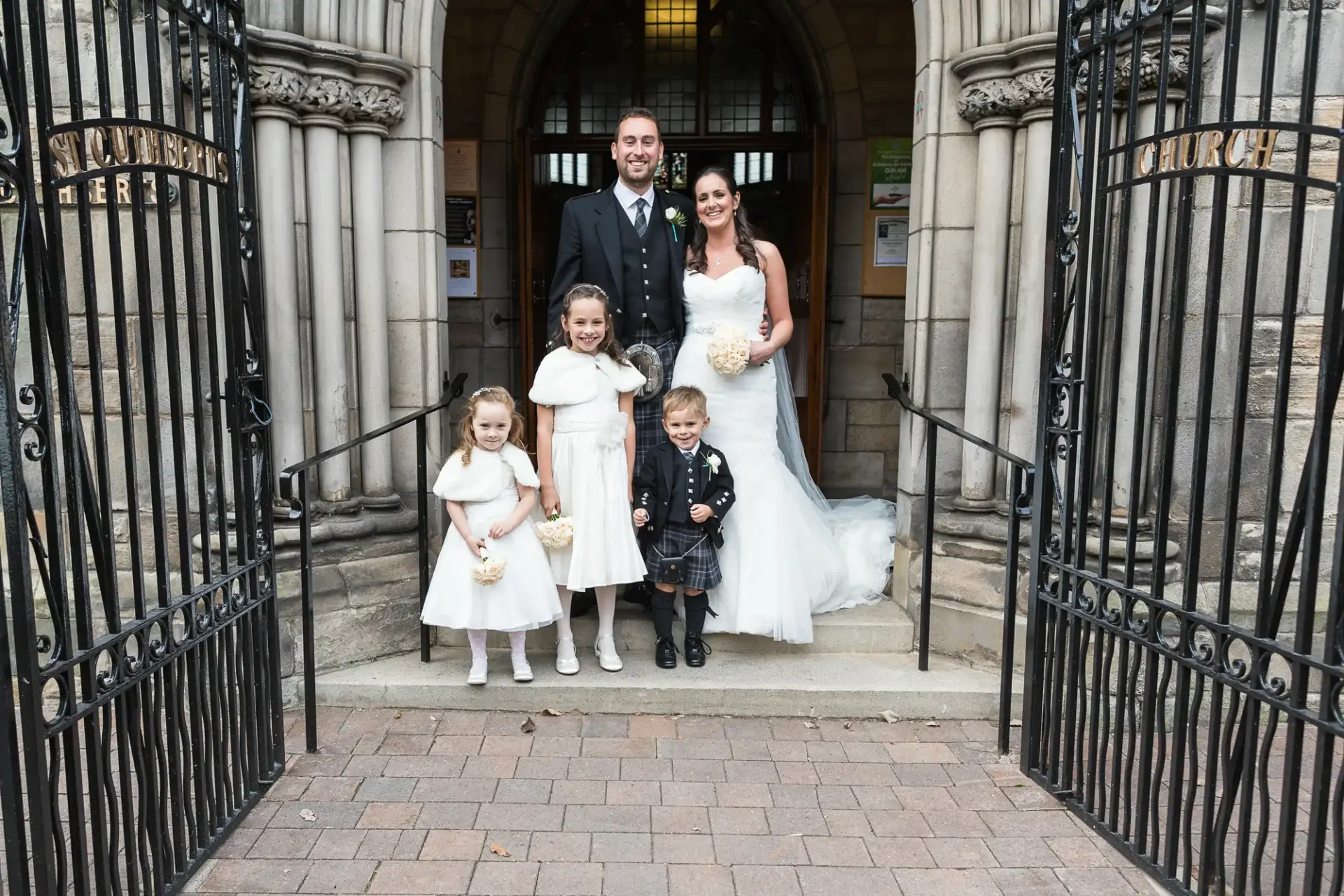 A bride and groom stand smiling with two flower girls and a ring bearer in front of a church entrance, with open wrought-iron gates.