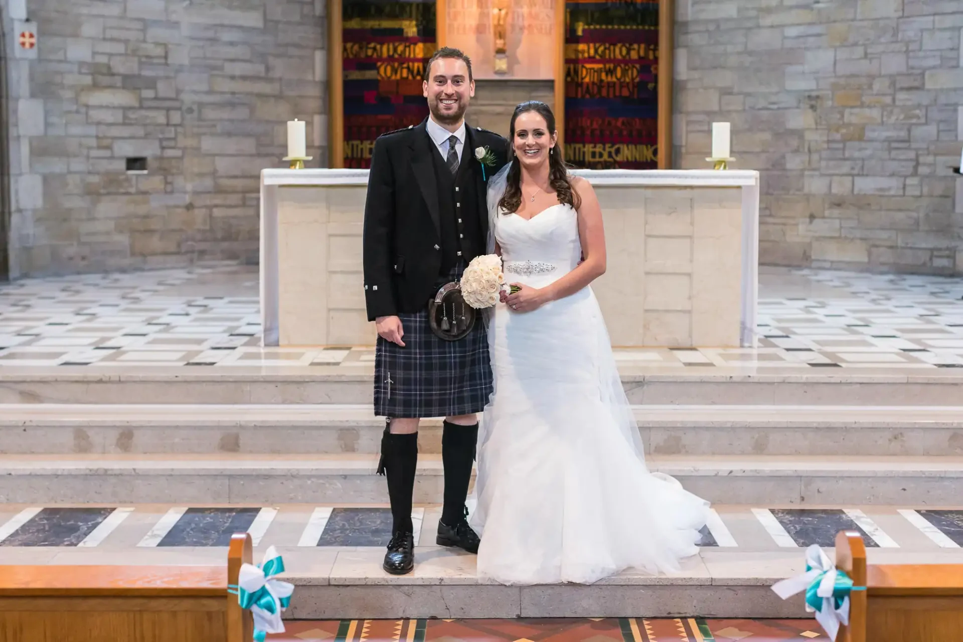 A bride and groom smiling, standing in a church; the groom wears a kilt and the bride is in a white gown, holding a bouquet.