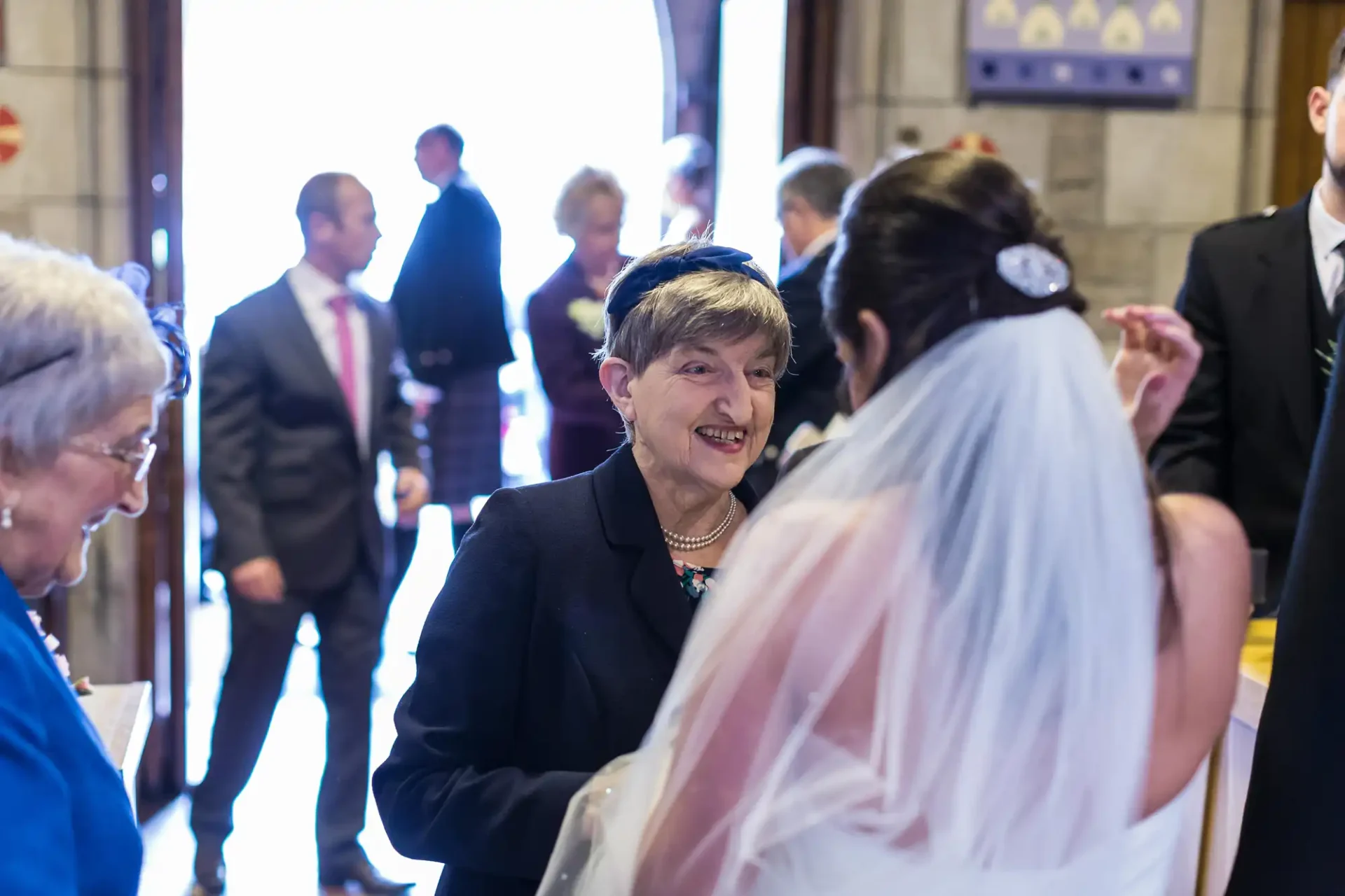 An elderly woman smiling joyfully at a bride in a veil inside a sunlit church, with other guests blurred in the background.