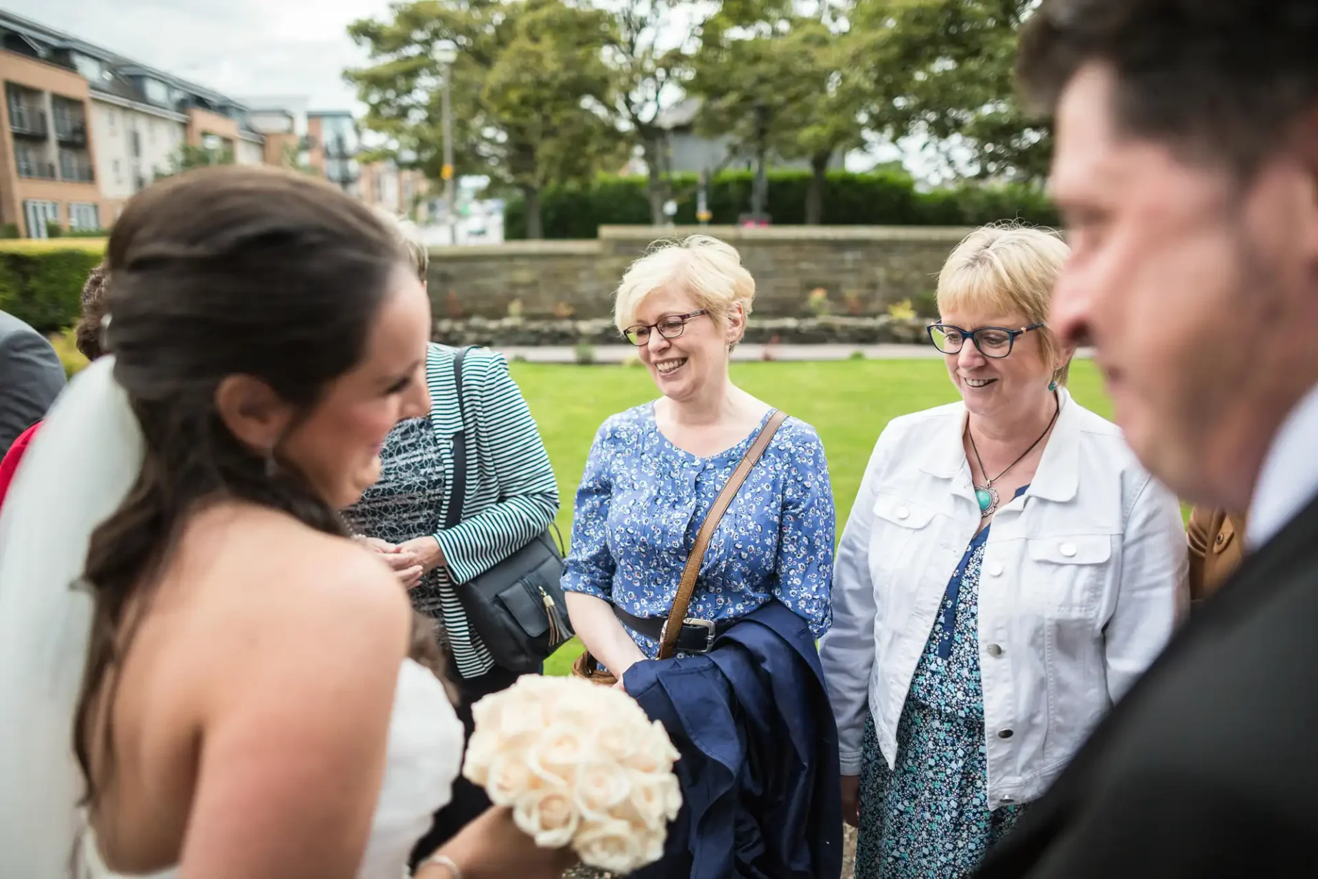 A bride holding a bouquet laughs joyfully with guests during a wedding reception outdoors.