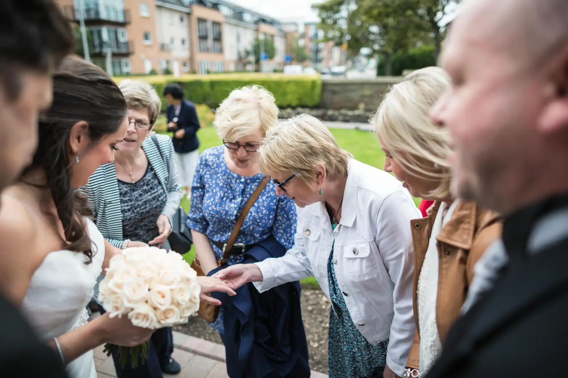 A bride shows her bouquet to a group of admiring guests at an outdoor wedding function.