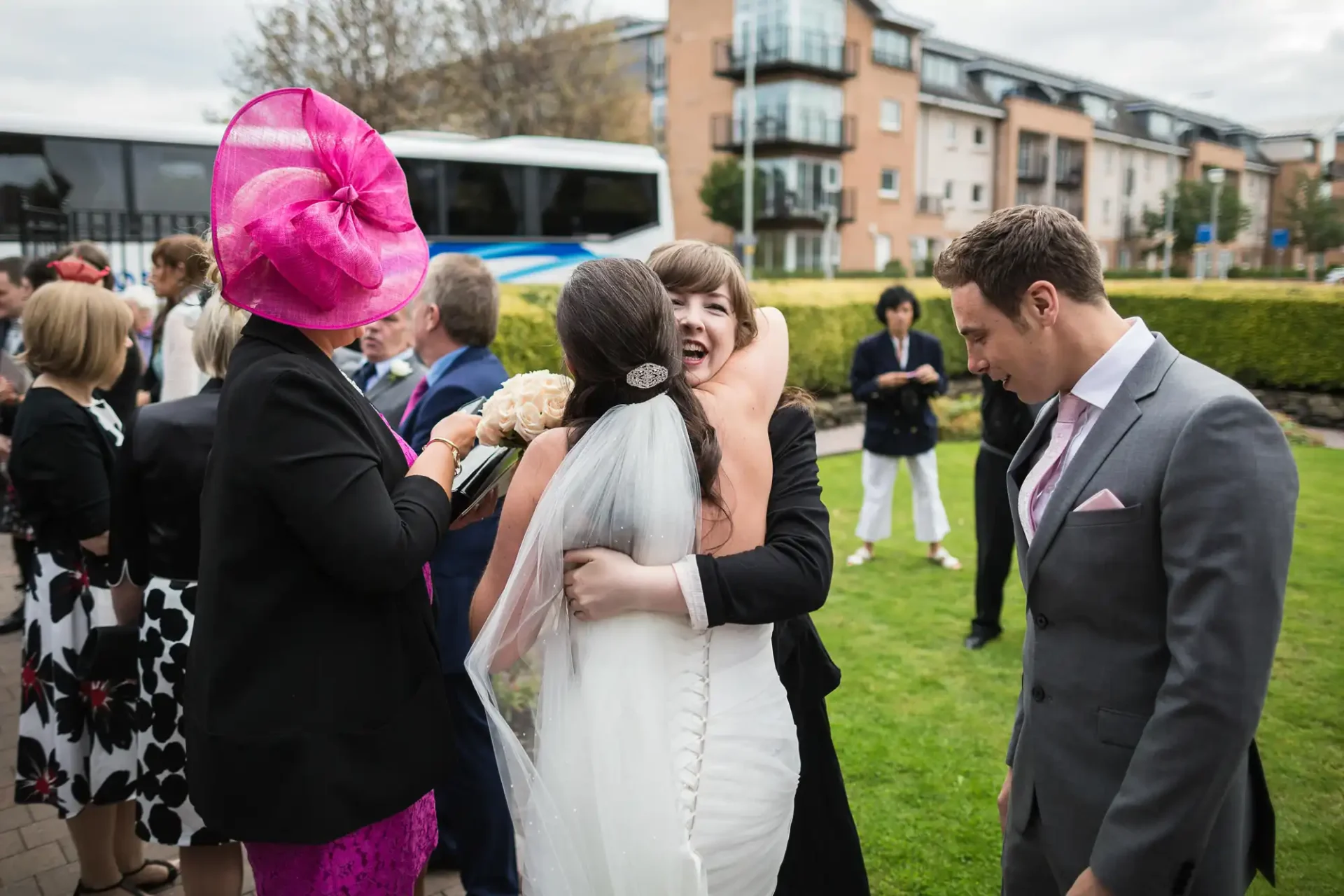 A bride in a white dress embraces a woman in a pink hat while others, including a man in a suit, look on at an outdoor gathering.